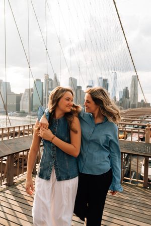Two friends share a laugh while walking on the Brooklyn Bridge with the Manhattan skyline in the background.