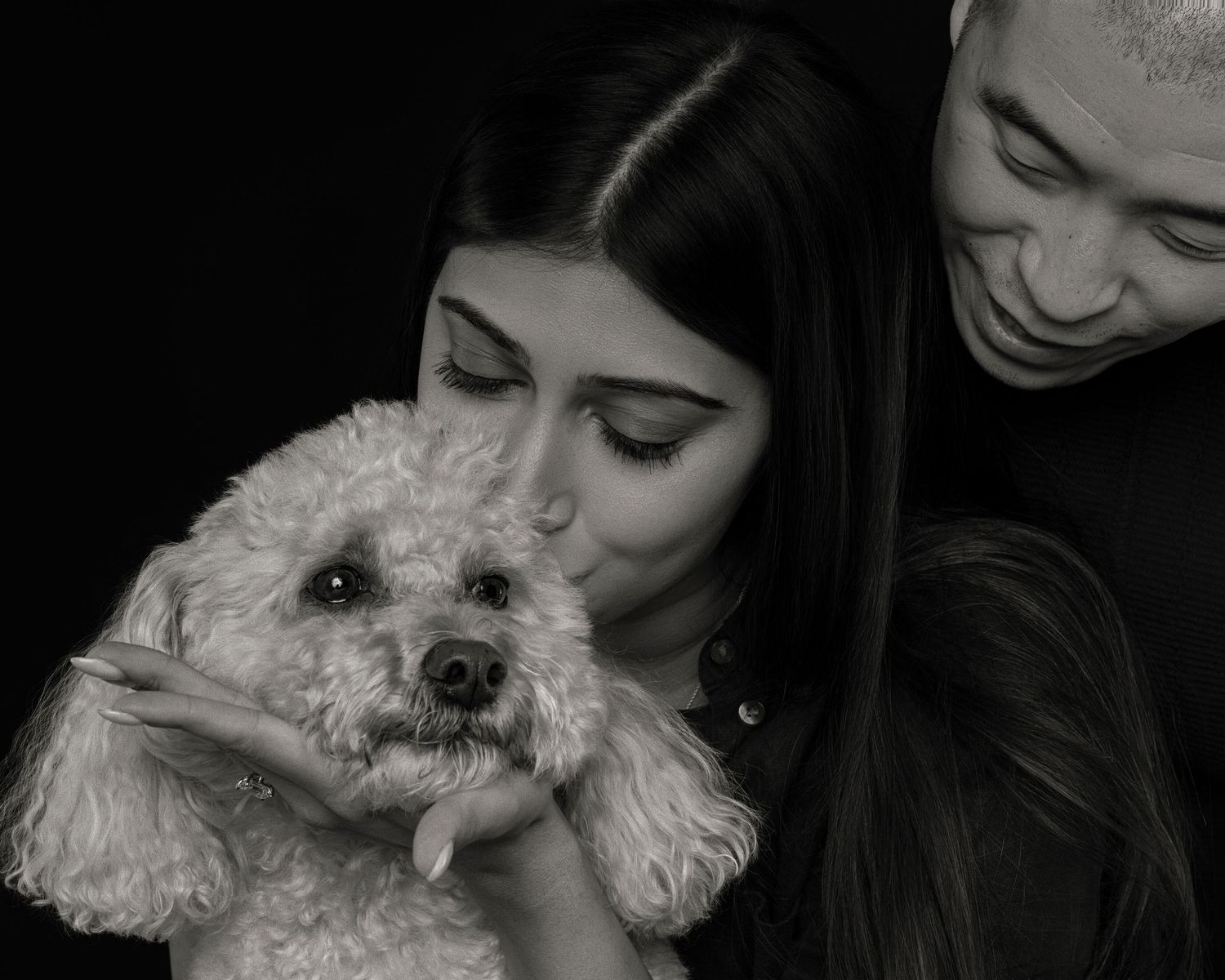 Black and white photo of a couple lovingly holding and kissing a fluffy white poodle dog.