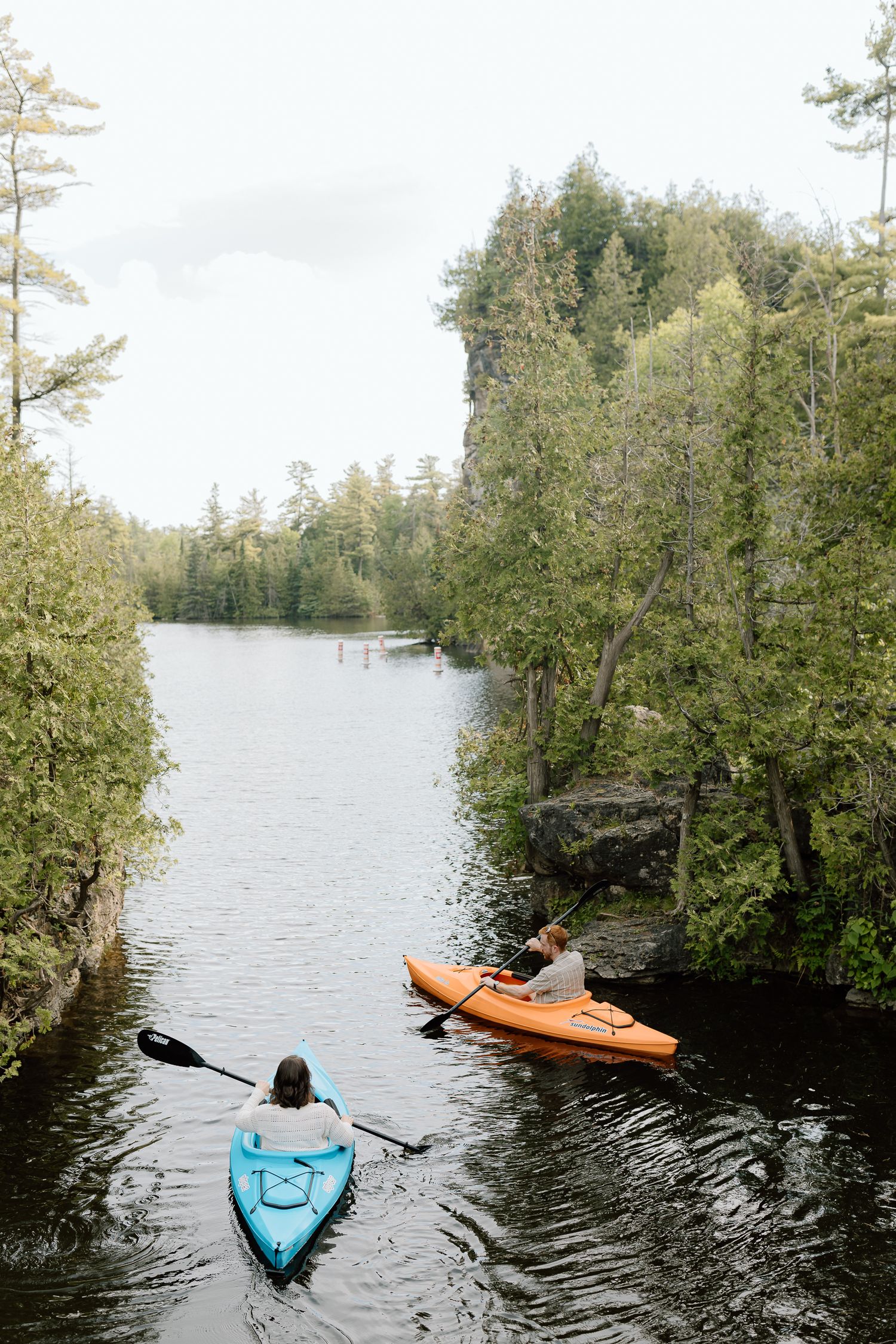 Two kayakers paddling blue and orange kayaks through a calm river surrounded by lush green trees and rocky cliffs.