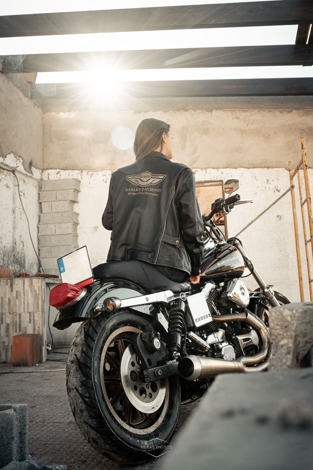 Woman in Harley-Davidson leather jacket sitting on a classic motorcycle in a rustic garage with sunlight.