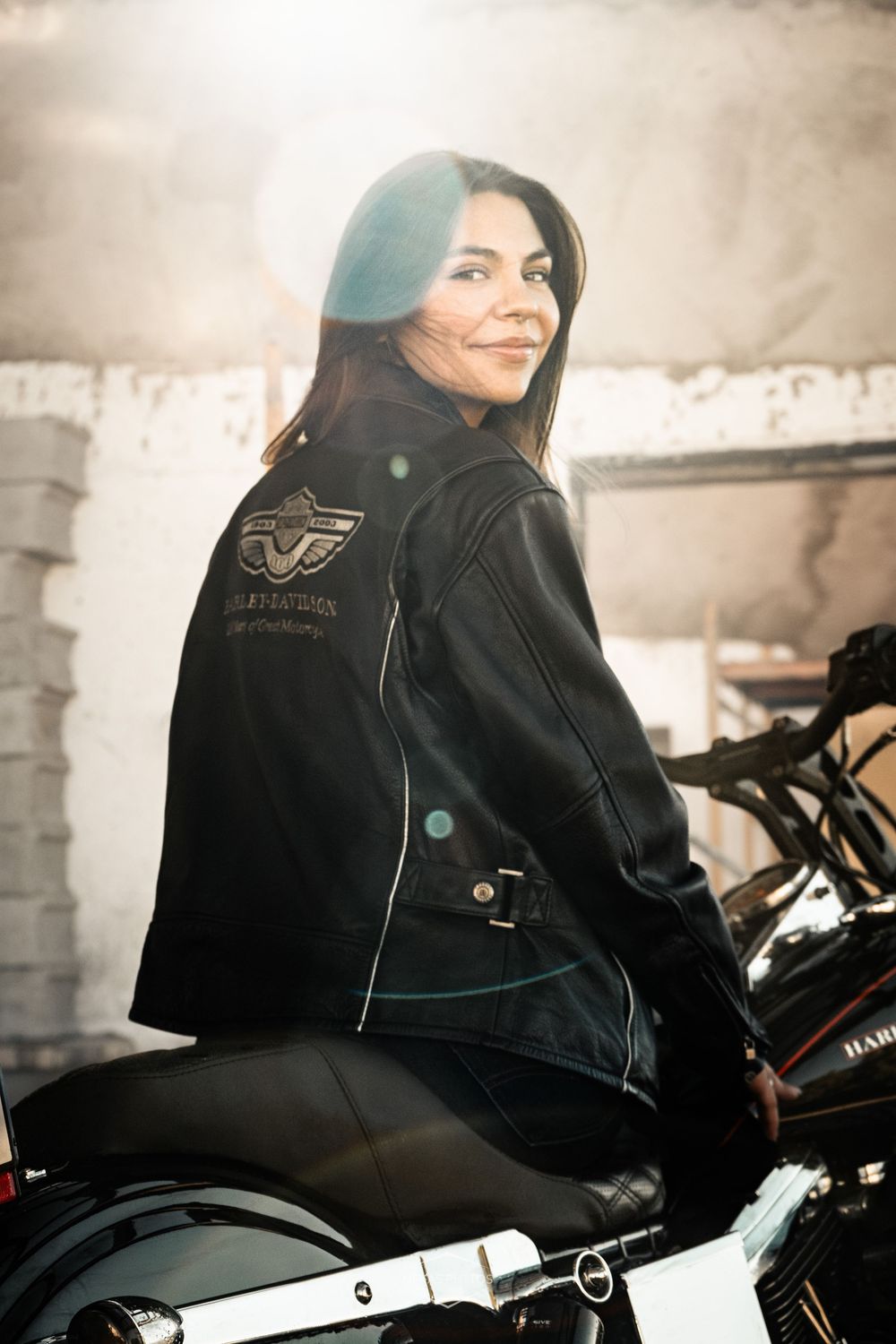 Woman in Harley-Davidson leather jacket sitting on a motorcycle, smiling over her shoulder in a garage.