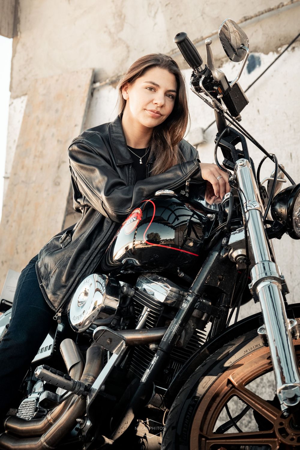 Young woman in black leather jacket sitting on a classic black motorcycle in an urban setting.
