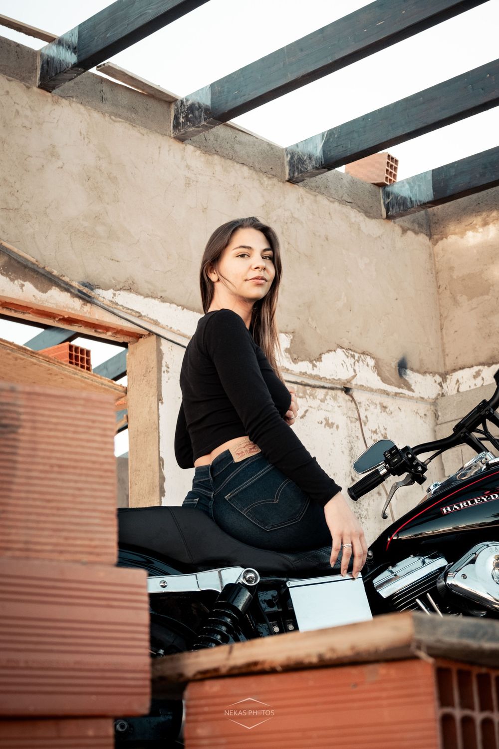 Woman in black outfit sitting on motorcycle in urban setting with weathered walls and exposed beams.
