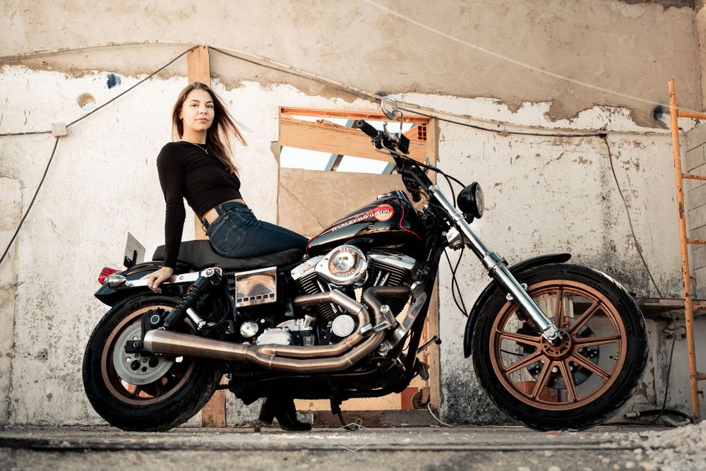 Young woman in black outfit posing on a classic Harley-Davidson chopper motorcycle against a rustic wall.
