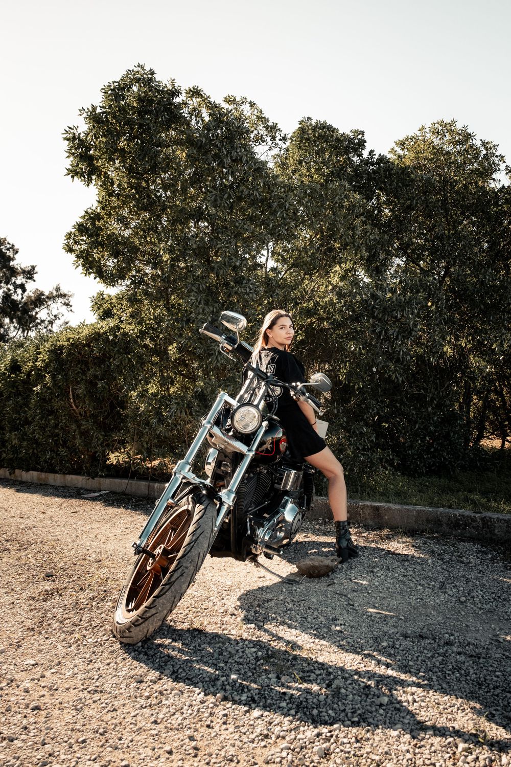 Woman in black outfit posing confidently on a classic motorcycle on a gravel path surrounded by lush green trees.