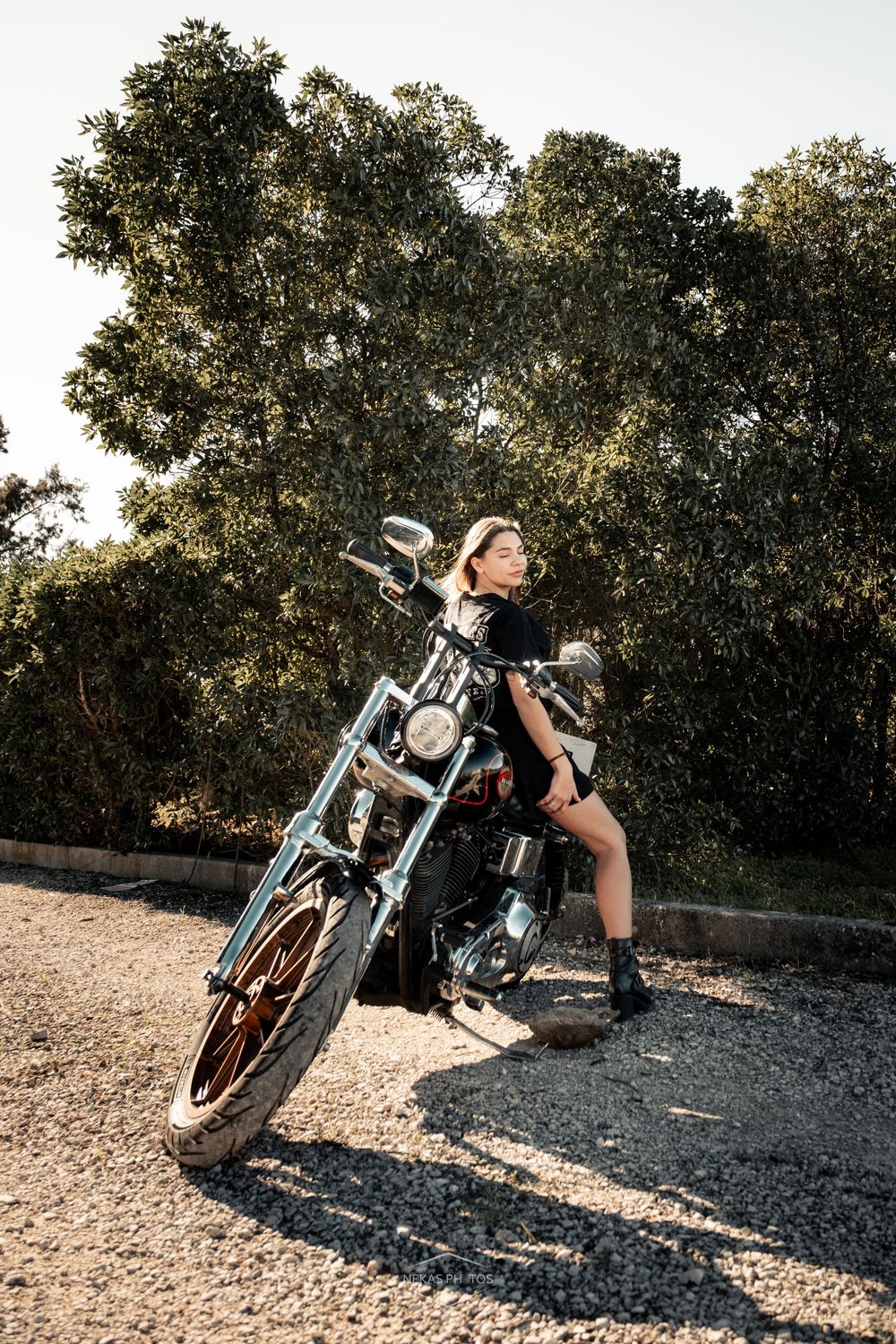 Young woman in black outfit posing on a classic chrome motorcycle on a gravel path surrounded by lush green trees.