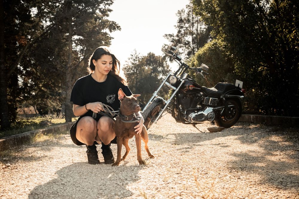 Woman crouching with brown dog on gravel path, motorcycle parked among trees in background.