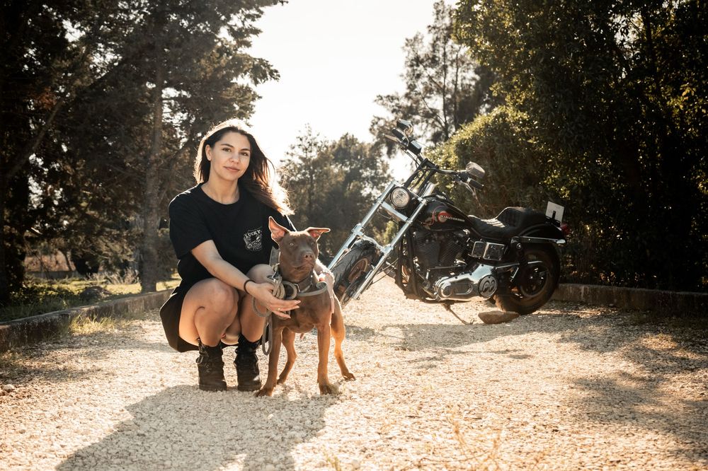 Woman crouching with a brown dog on a gravel path, with a motorcycle parked in the background among trees.