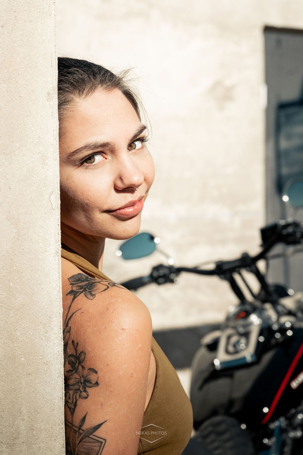 Young woman with floral shoulder tattoo leaning against wall with motorcycle in background.