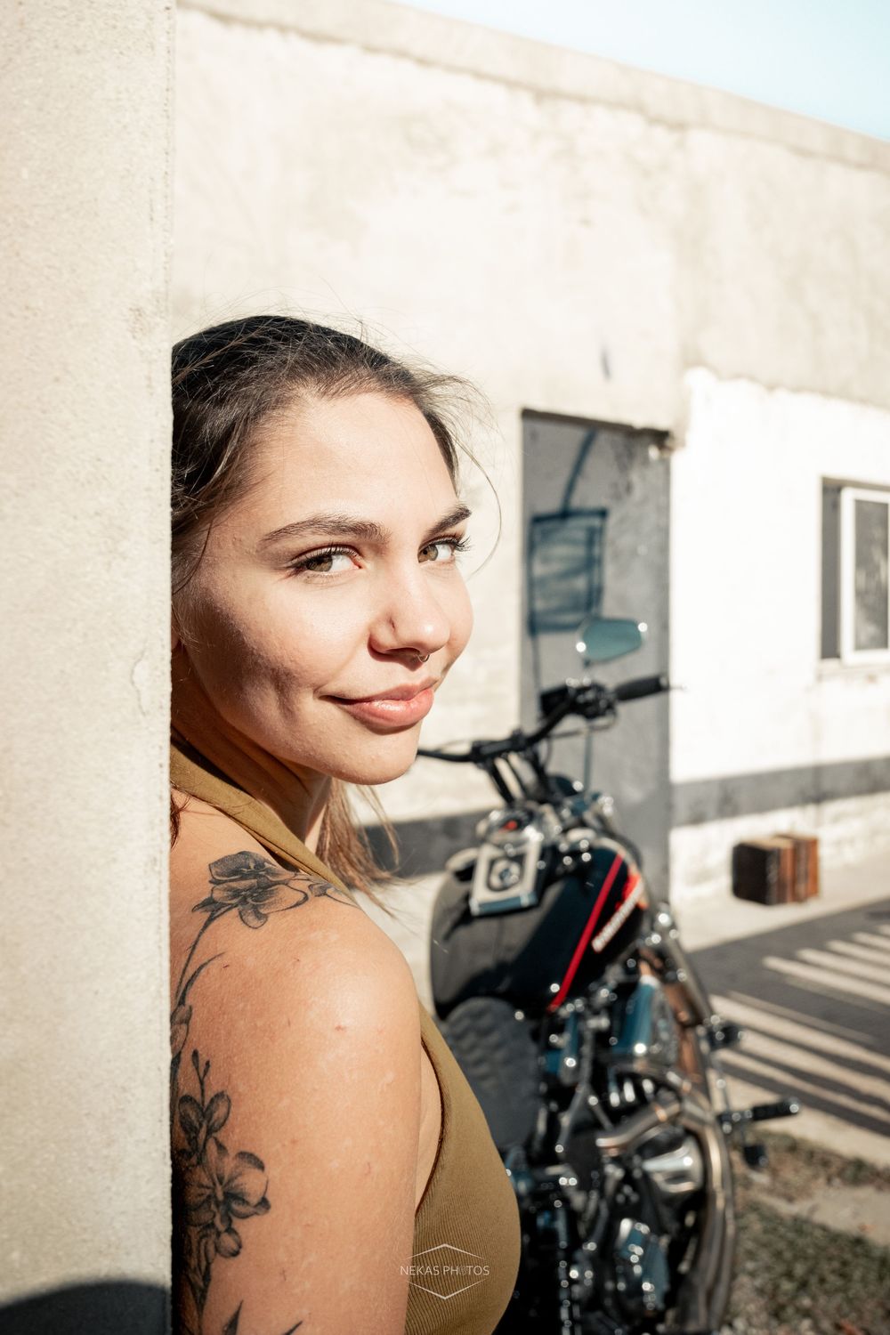 A tattooed woman leans against a wall smiling, with a custom motorcycle parked in the background on a sunny day.