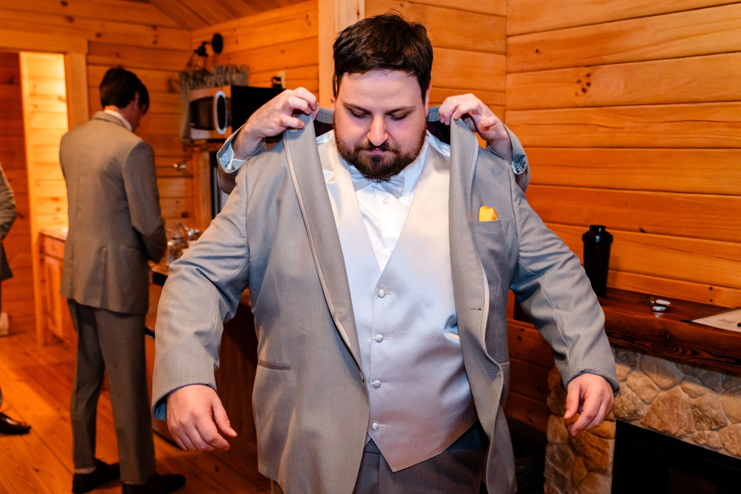 A groom puts on his gray wedding suit jacket in a rustic log cabin with a groomsman visible in background.