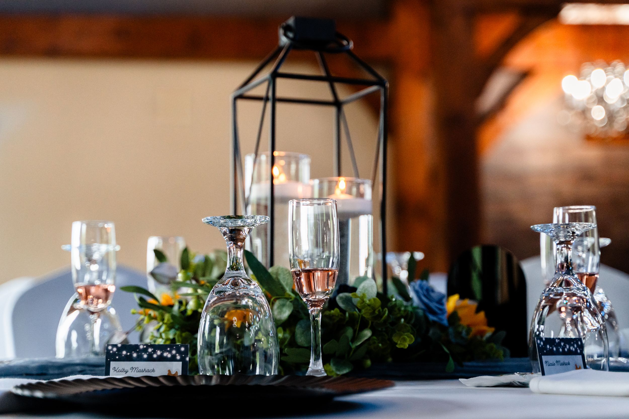 Elegant wedding reception table with a black lantern centerpiece, candles, champagne flutes, and floral greenery.