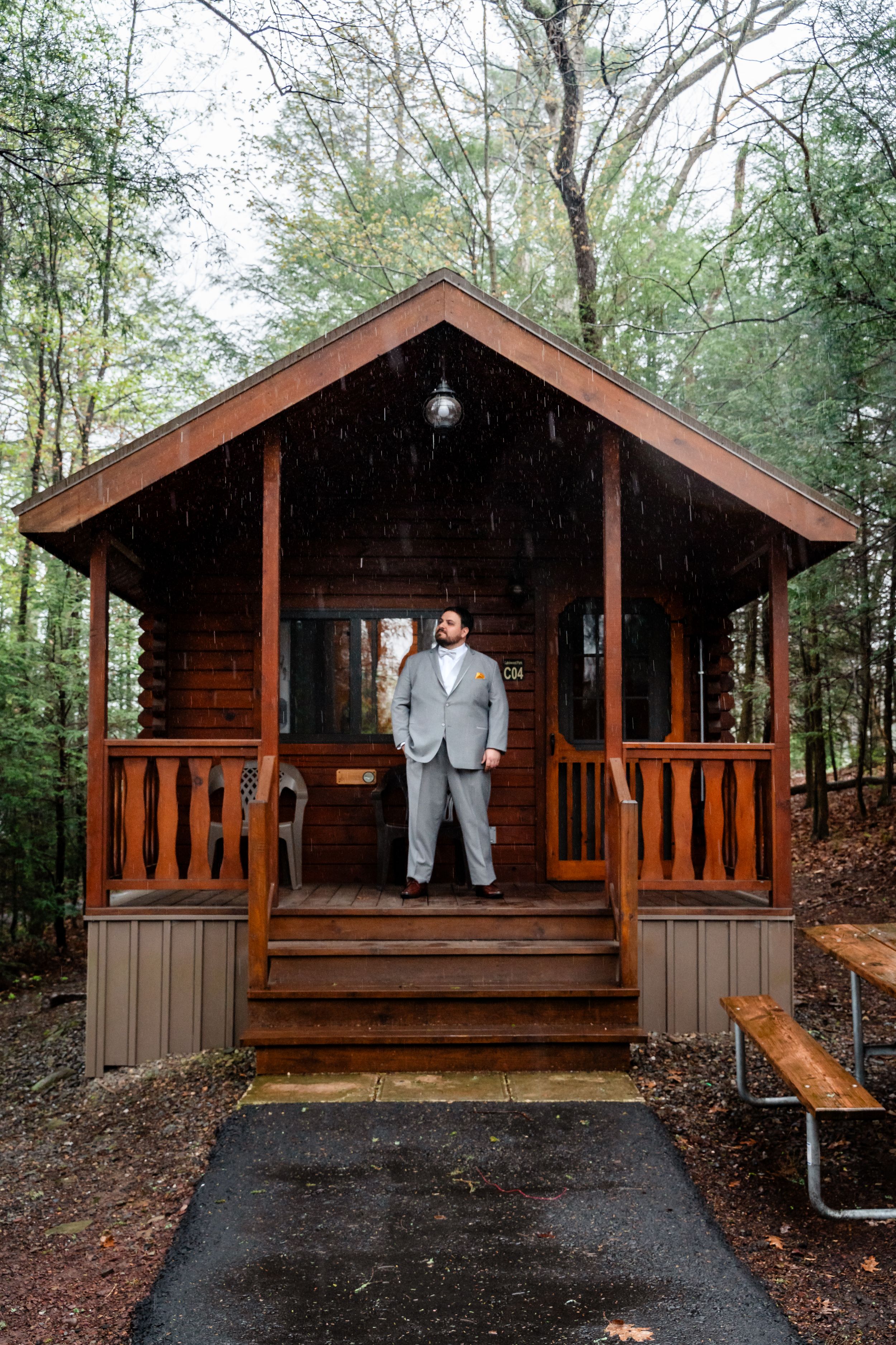 Groom in gray suit standing on wooden cabin porch in rainy forest setting.