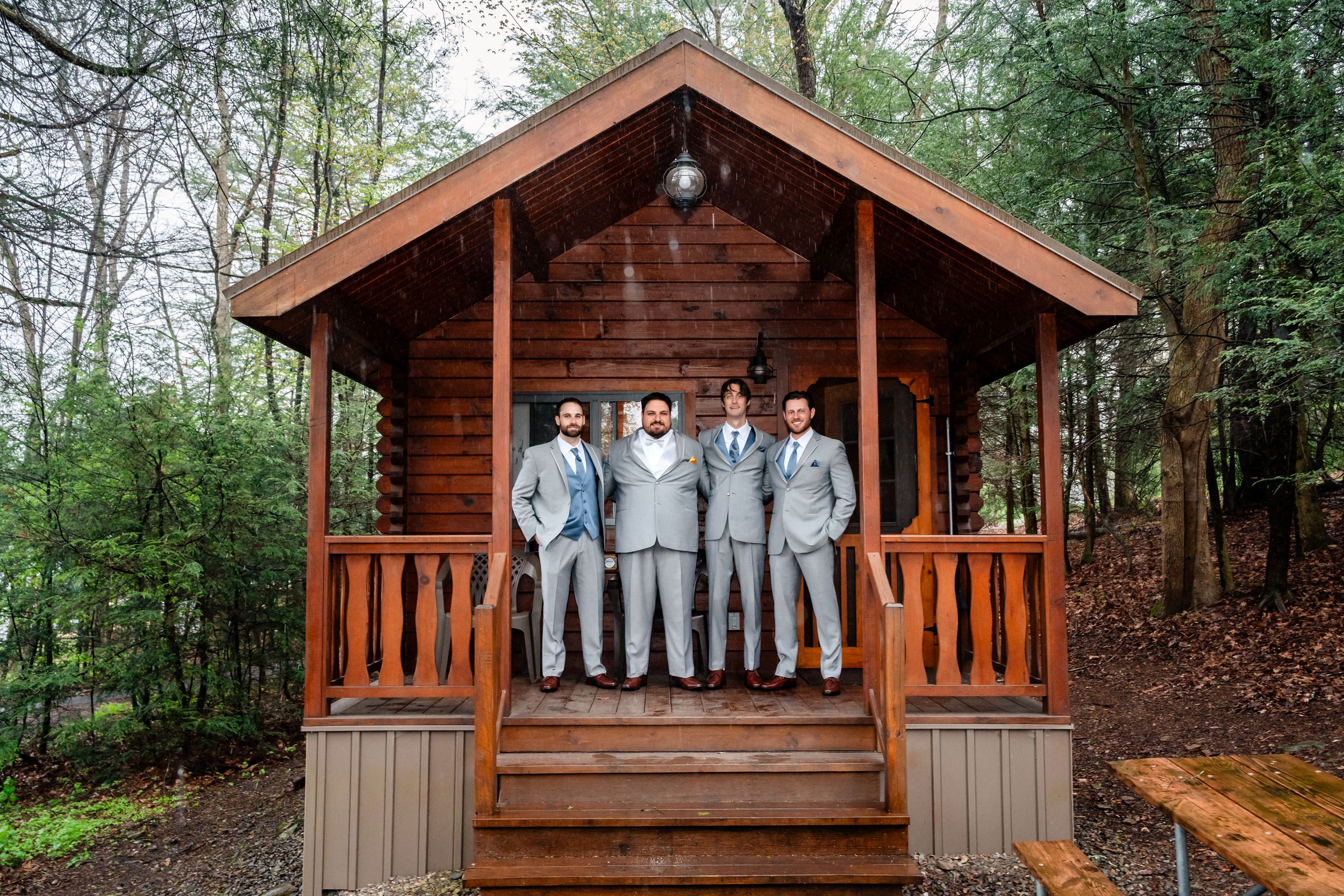 Groomsmen in gray suits posing on a rustic wooden cabin porch surrounded by lush forest trees.
