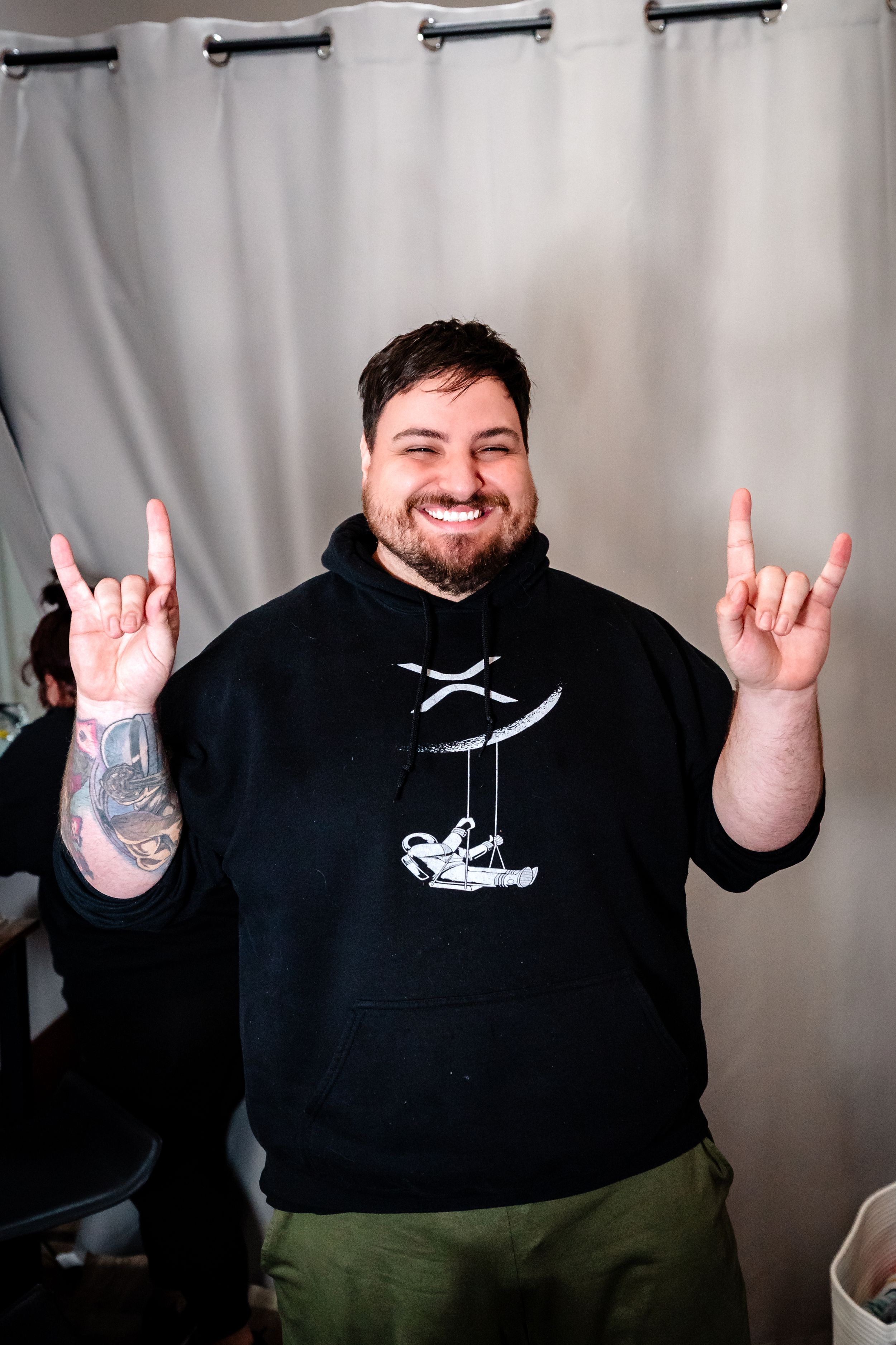 Smiling bearded man in black hoodie making rock on hand gestures with visible arm tattoo.