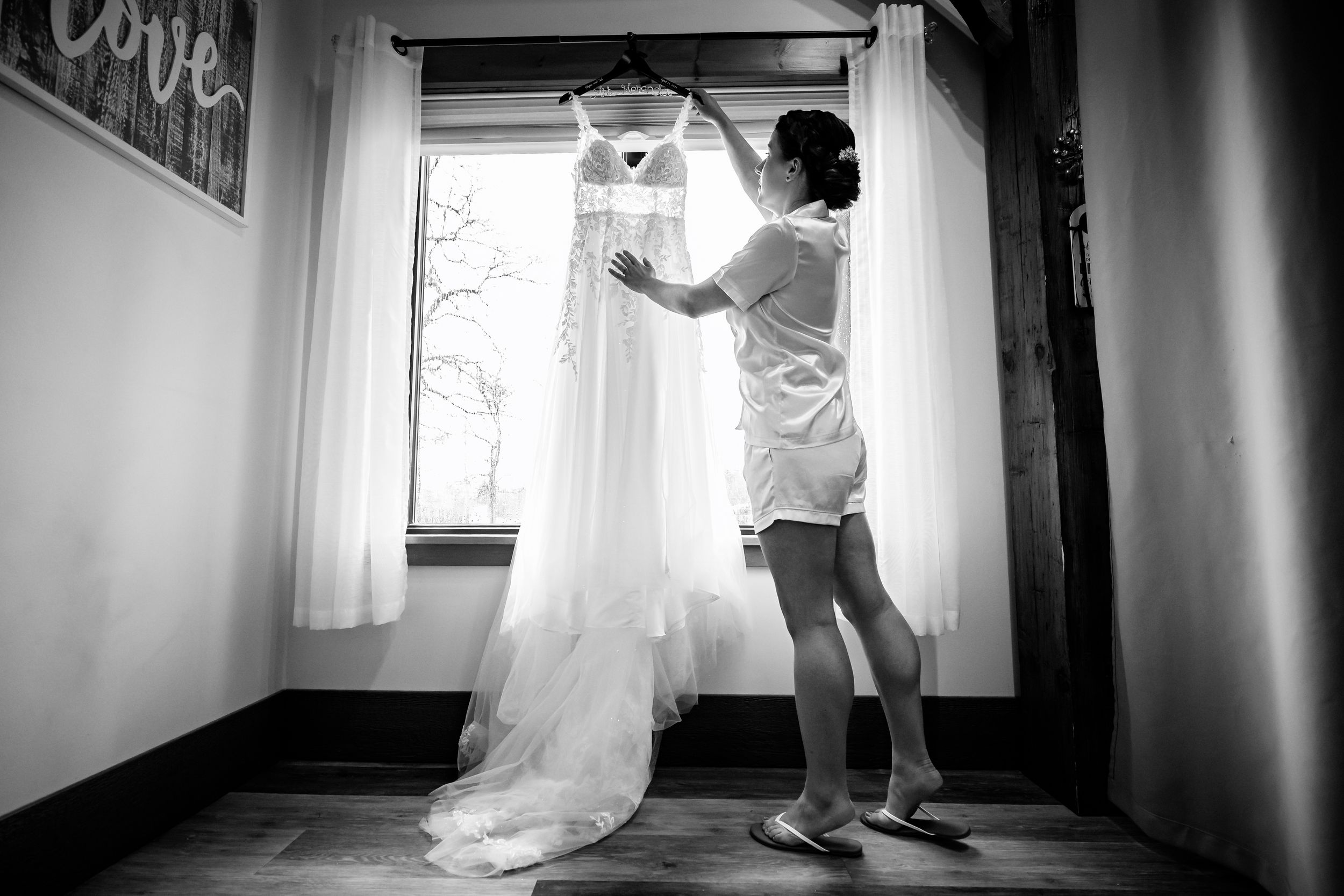 Black and white photo of a bride admiring her wedding gown hanging by a window in a rustic venue.