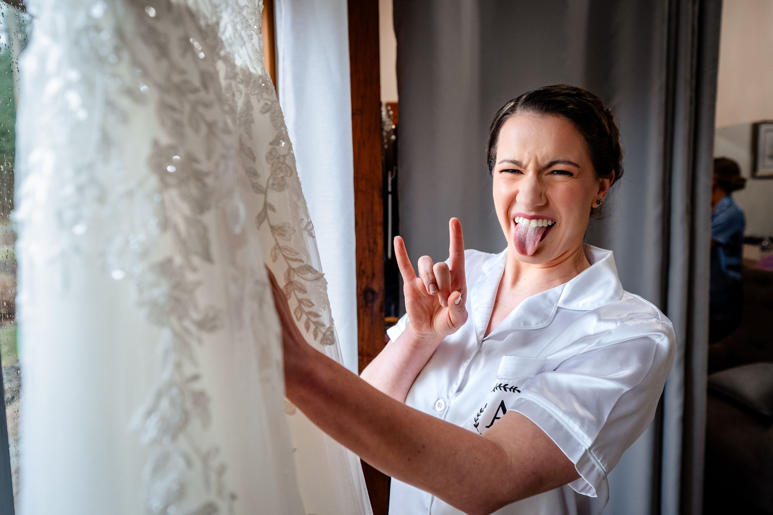 An excited bride-to-be makes a fun rock sign gesture while standing next to her lace wedding dress.