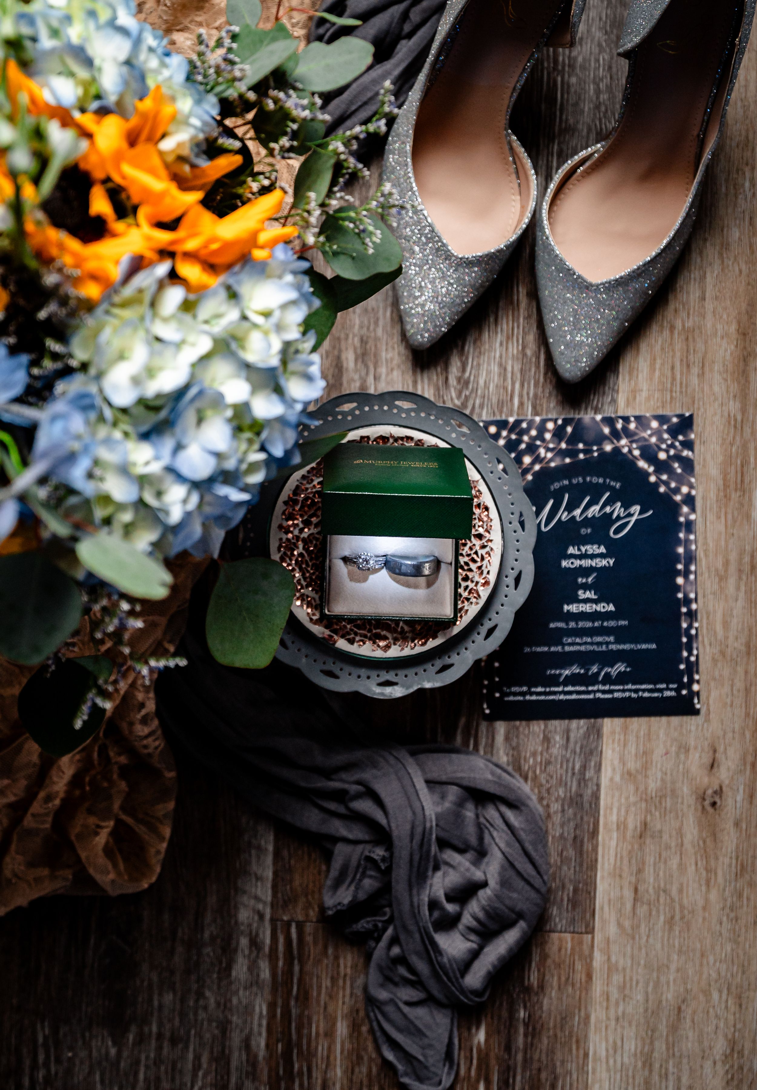 Wedding flat lay with floral bouquet, glitter heels, ring box, and navy invitation on rustic wood.