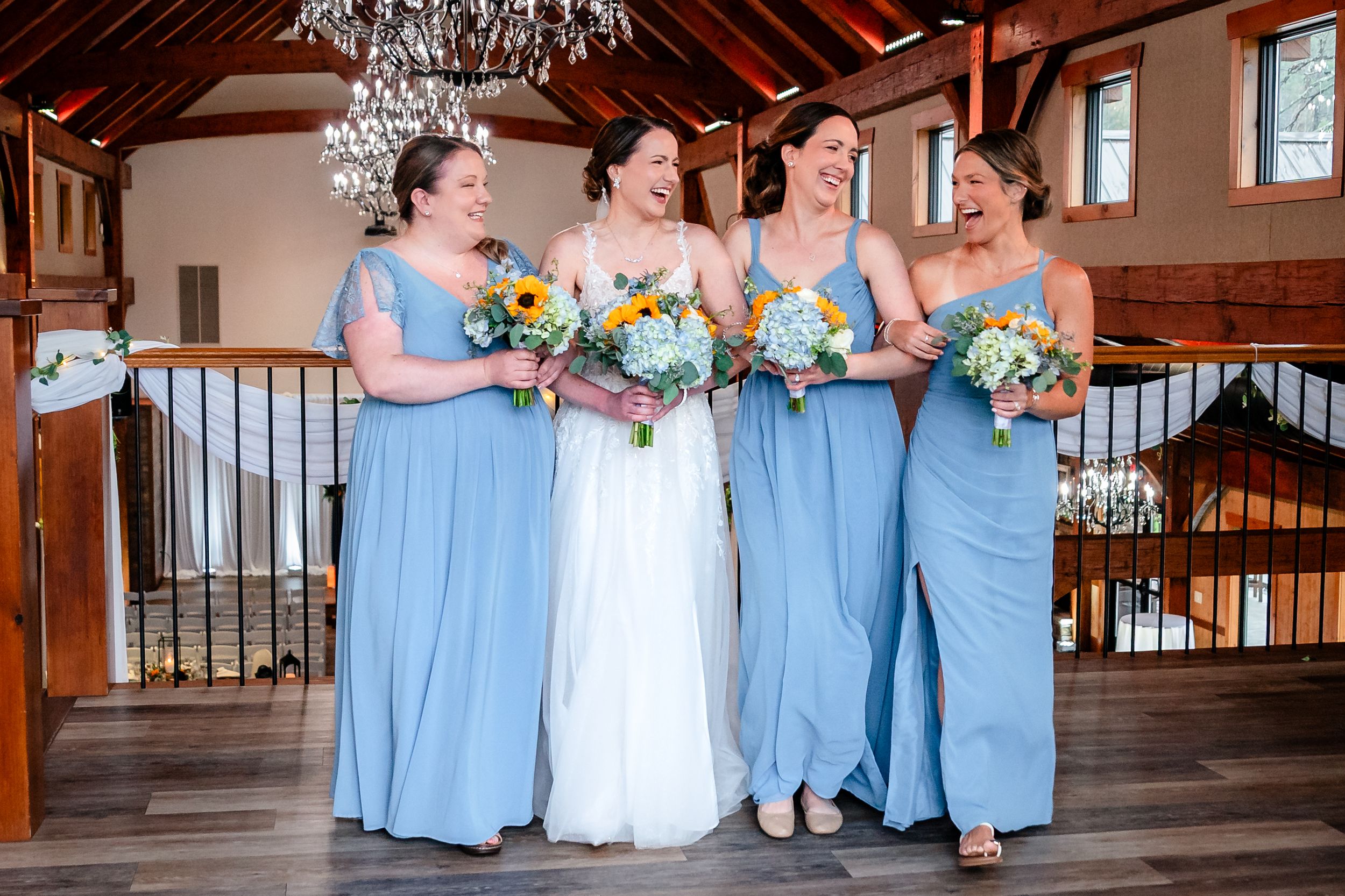 Bride and three bridesmaids in blue dresses laughing together inside a rustic wedding venue with chandelier.