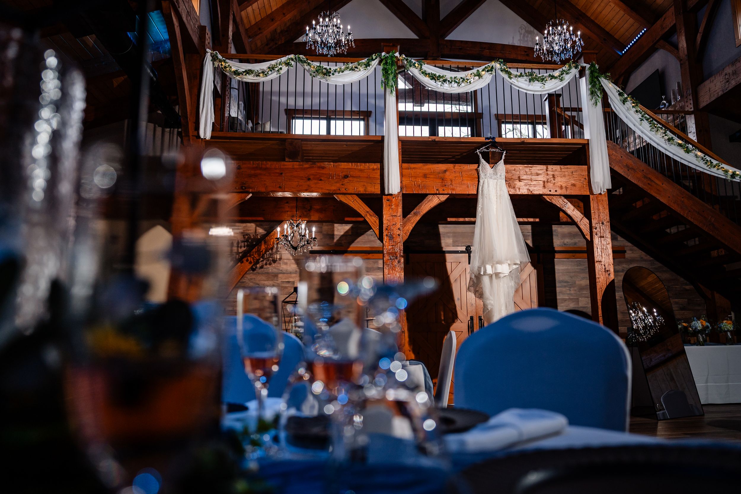 Elegant wedding dress displayed in a rustic barn venue with chandelier lighting and decorated tables.