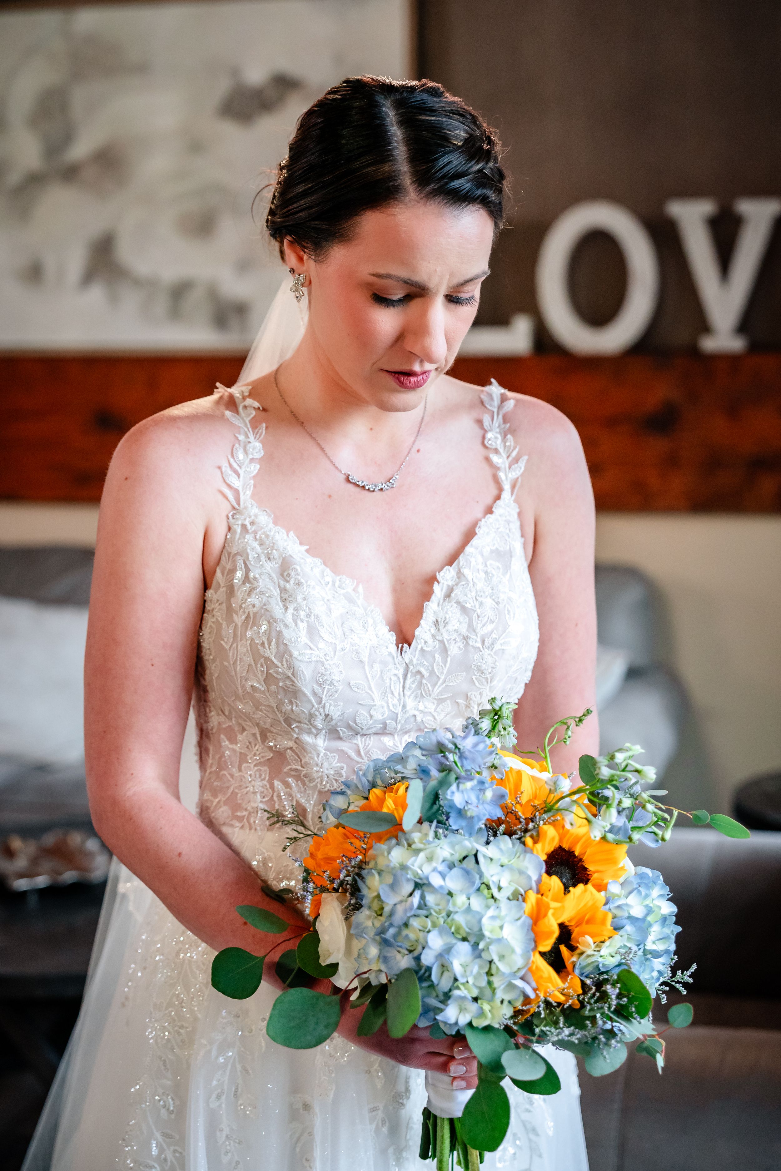 Bride in lace wedding gown holding blue and orange bouquet with sunflowers and hydrangeas, looking down lovingly.