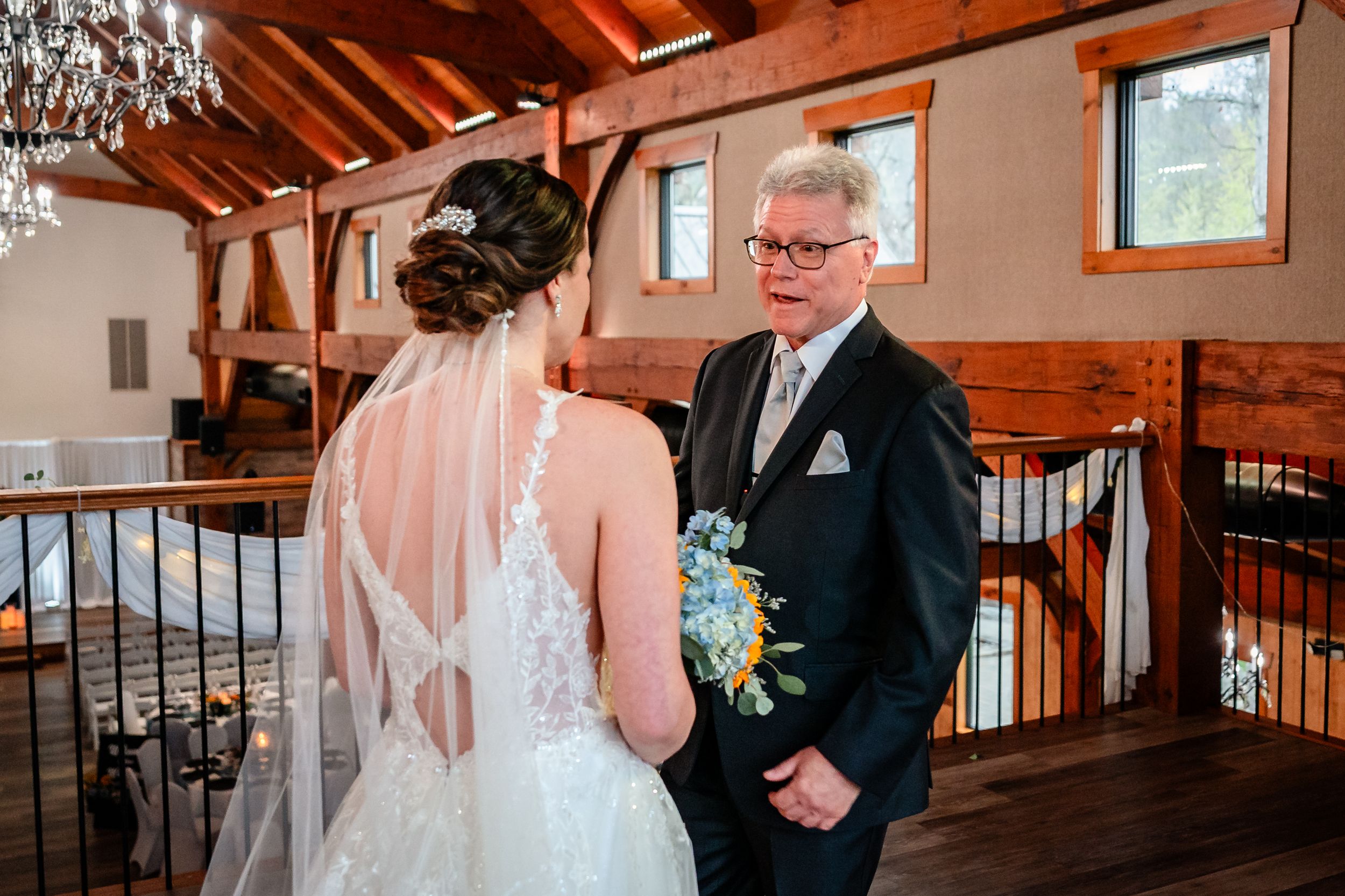 A bride in a backless lace wedding gown shares an emotional first look moment with her father in a rustic barn.