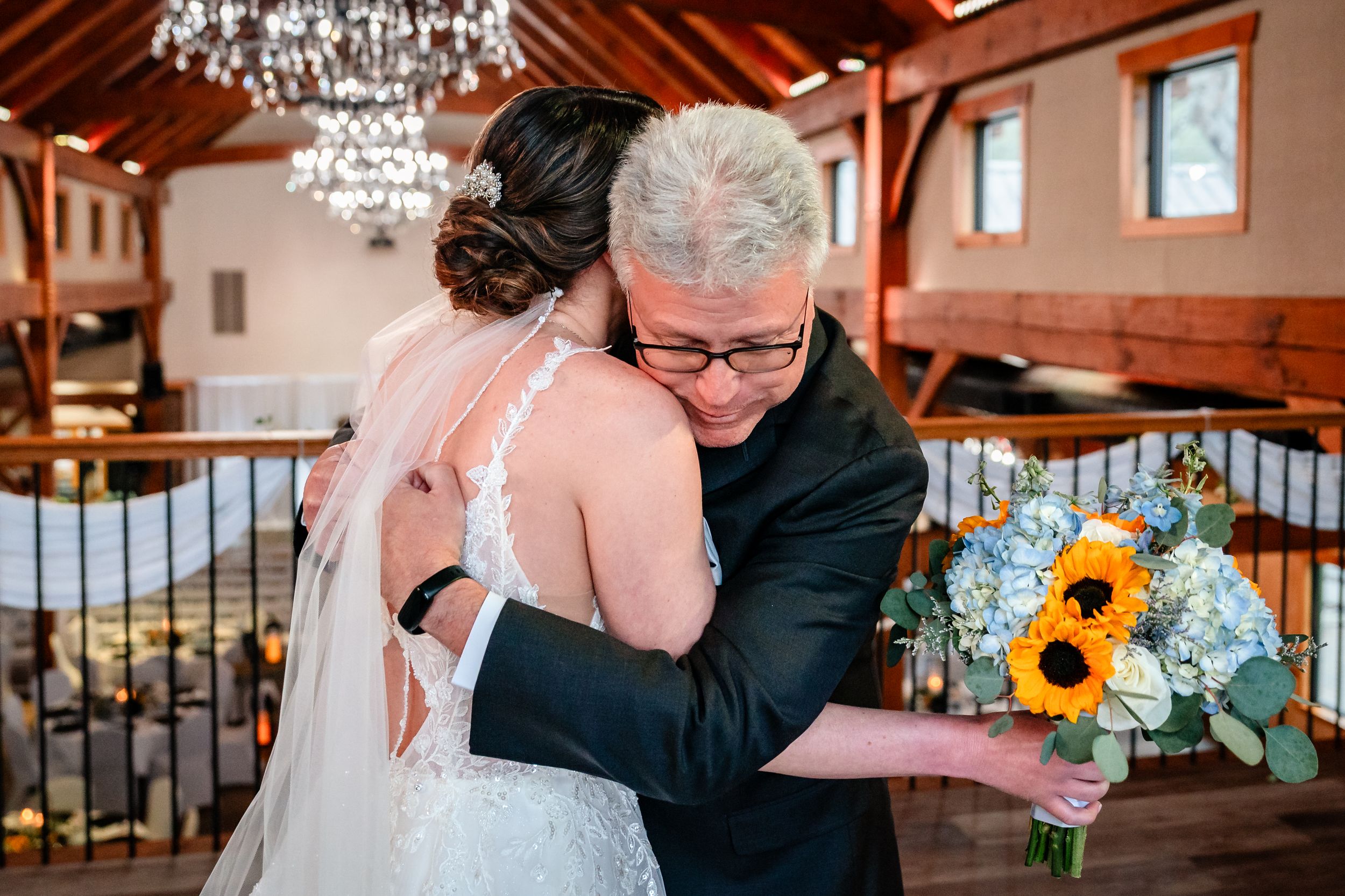 Bride receiving an emotional hug from older man holding sunflower bouquet in rustic wedding venue.