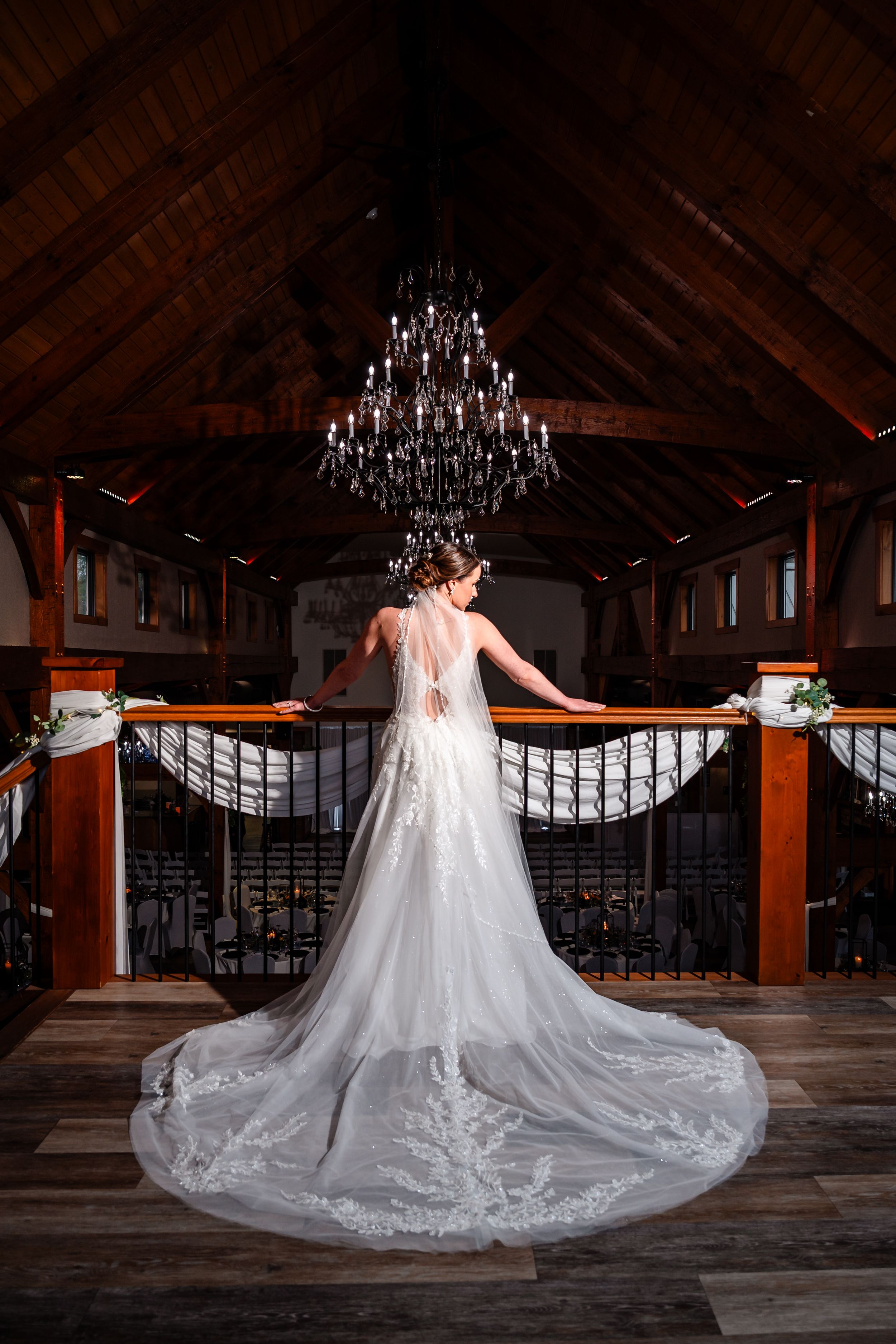 Bride in lace gown with long train stands at balcony railing beneath chandelier in elegant barn venue.