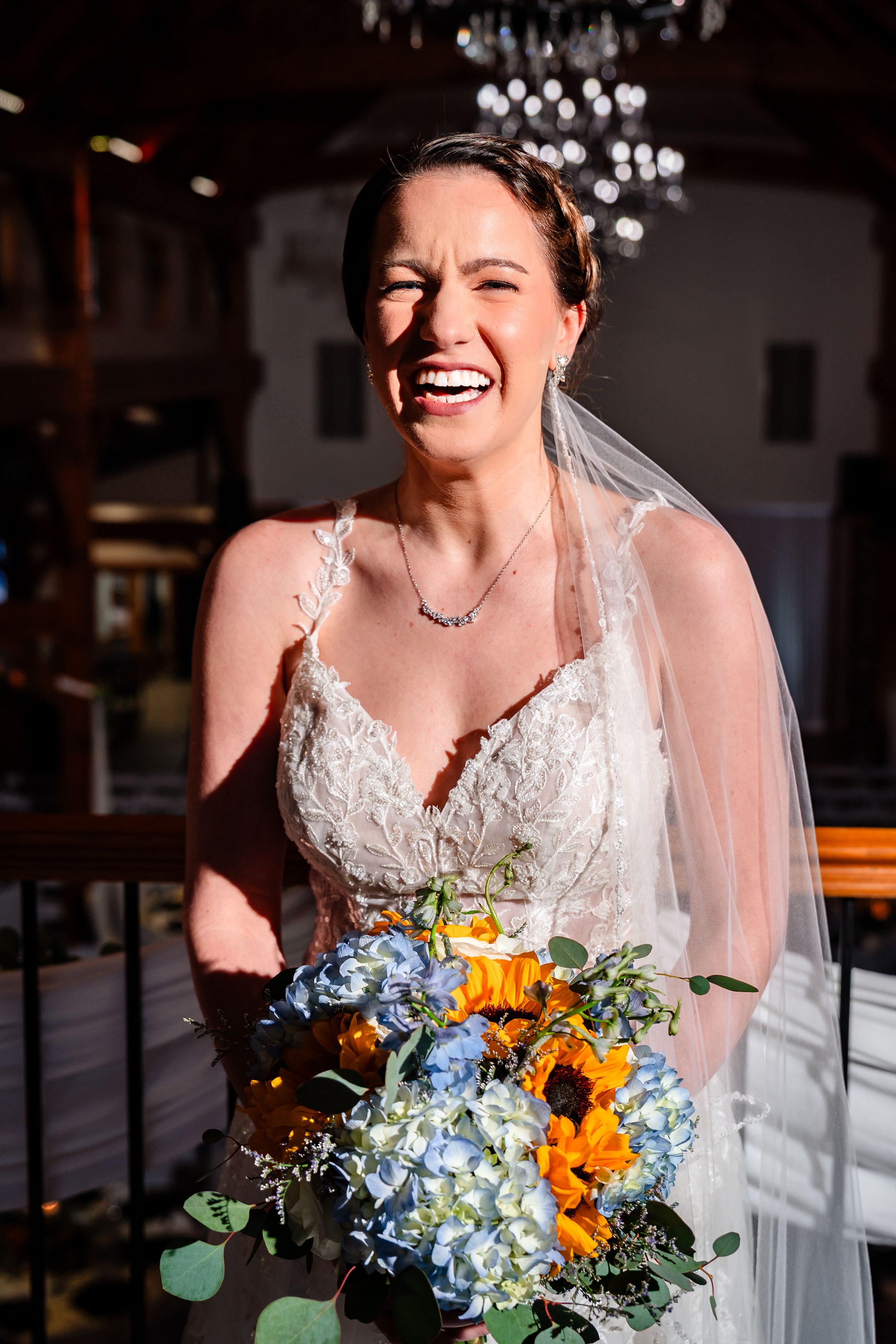 Radiant bride laughing in sunlight wearing lace gown and veil holding sunflower bouquet.