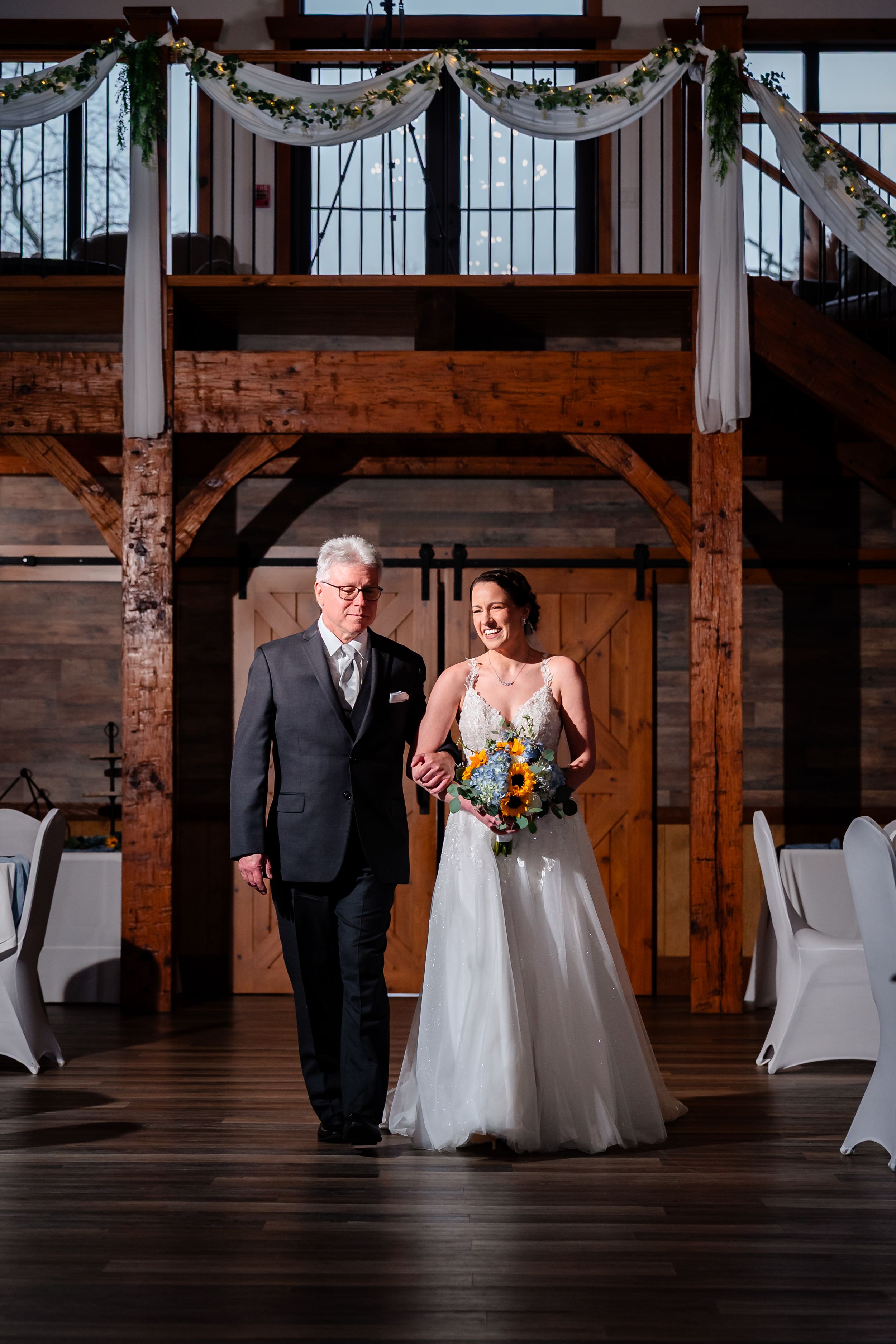Bride walks down aisle with escort in rustic barn wedding venue decorated with greenery and white chairs.