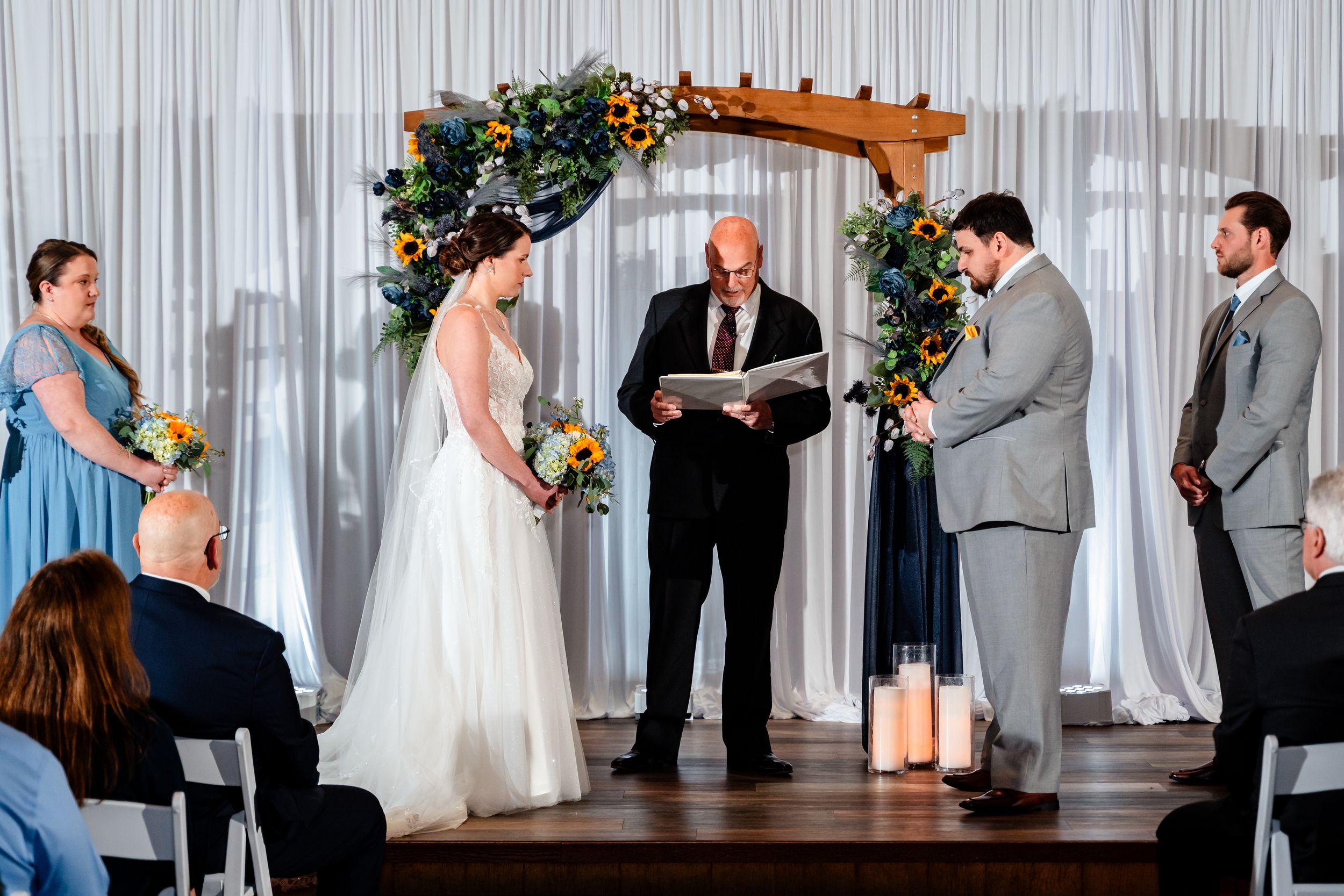 Bride and groom exchange vows under wooden arch with blue and yellow florals at indoor wedding ceremony.