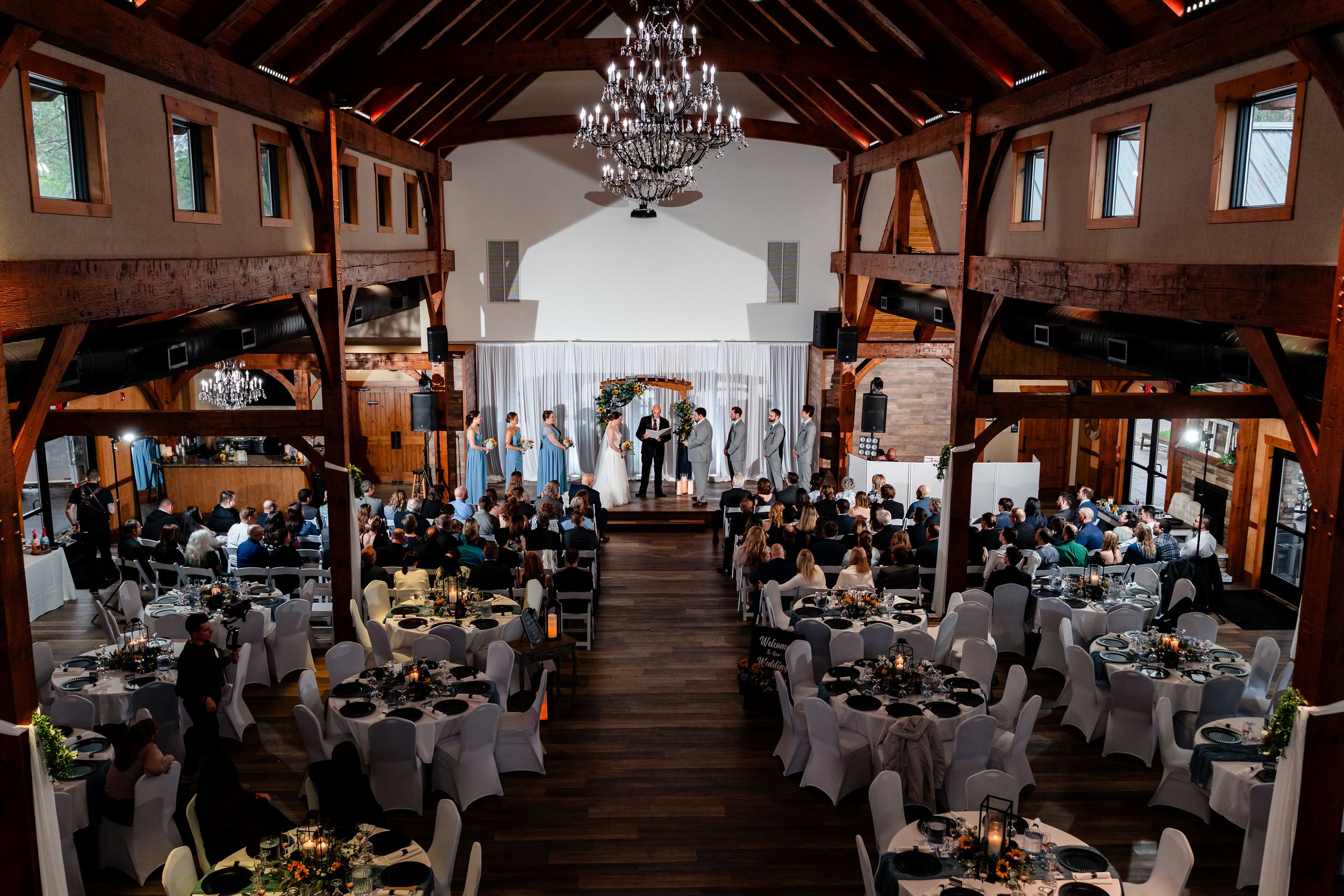 Aerial view of rustic barn wedding ceremony with guests seated at round tables under grand chandelier.