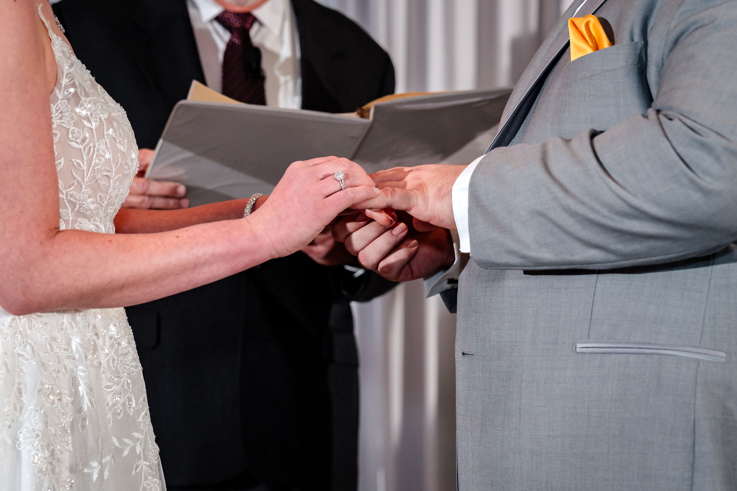 Couple exchanges wedding rings during ceremony while officiant holds book behind them.