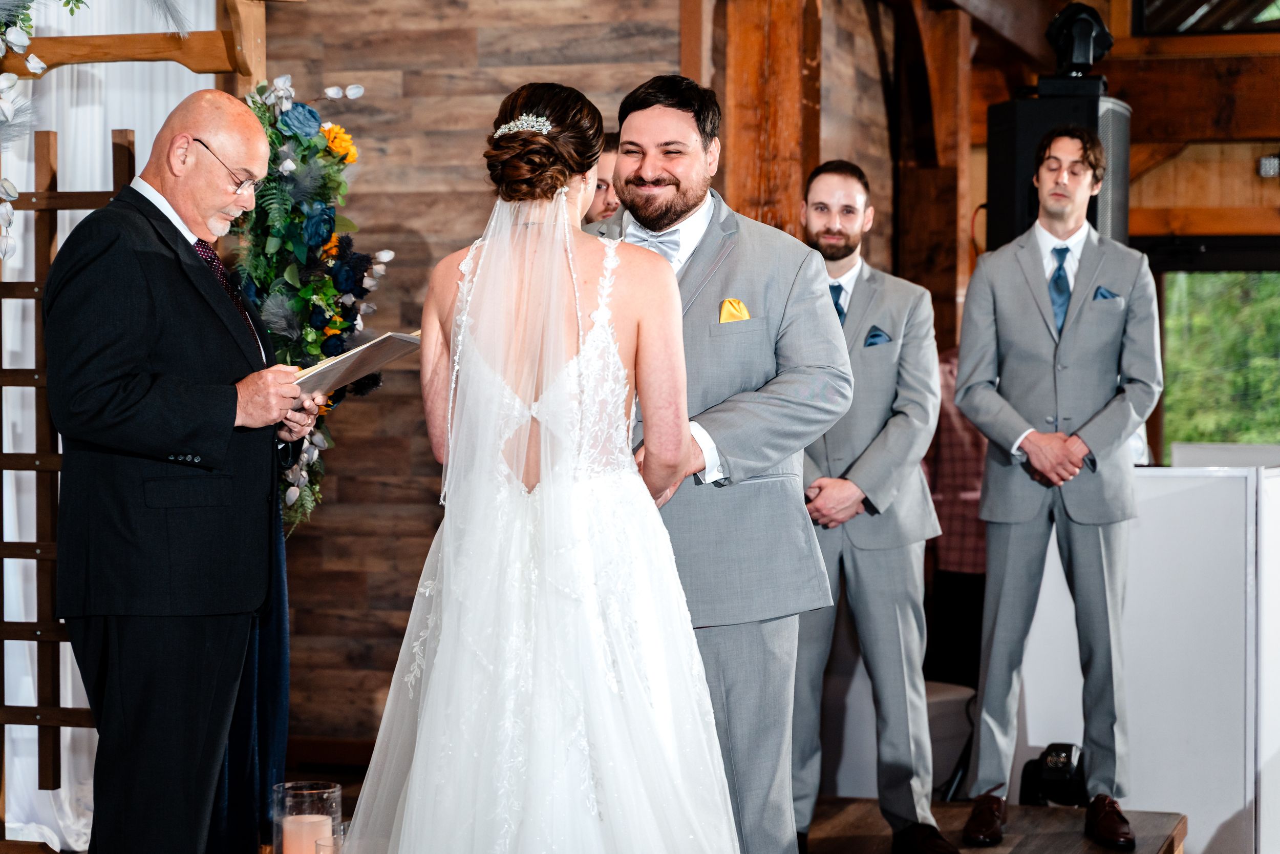 Groom smiling at bride during an indoor rustic barn wedding ceremony with groomsmen standing nearby.