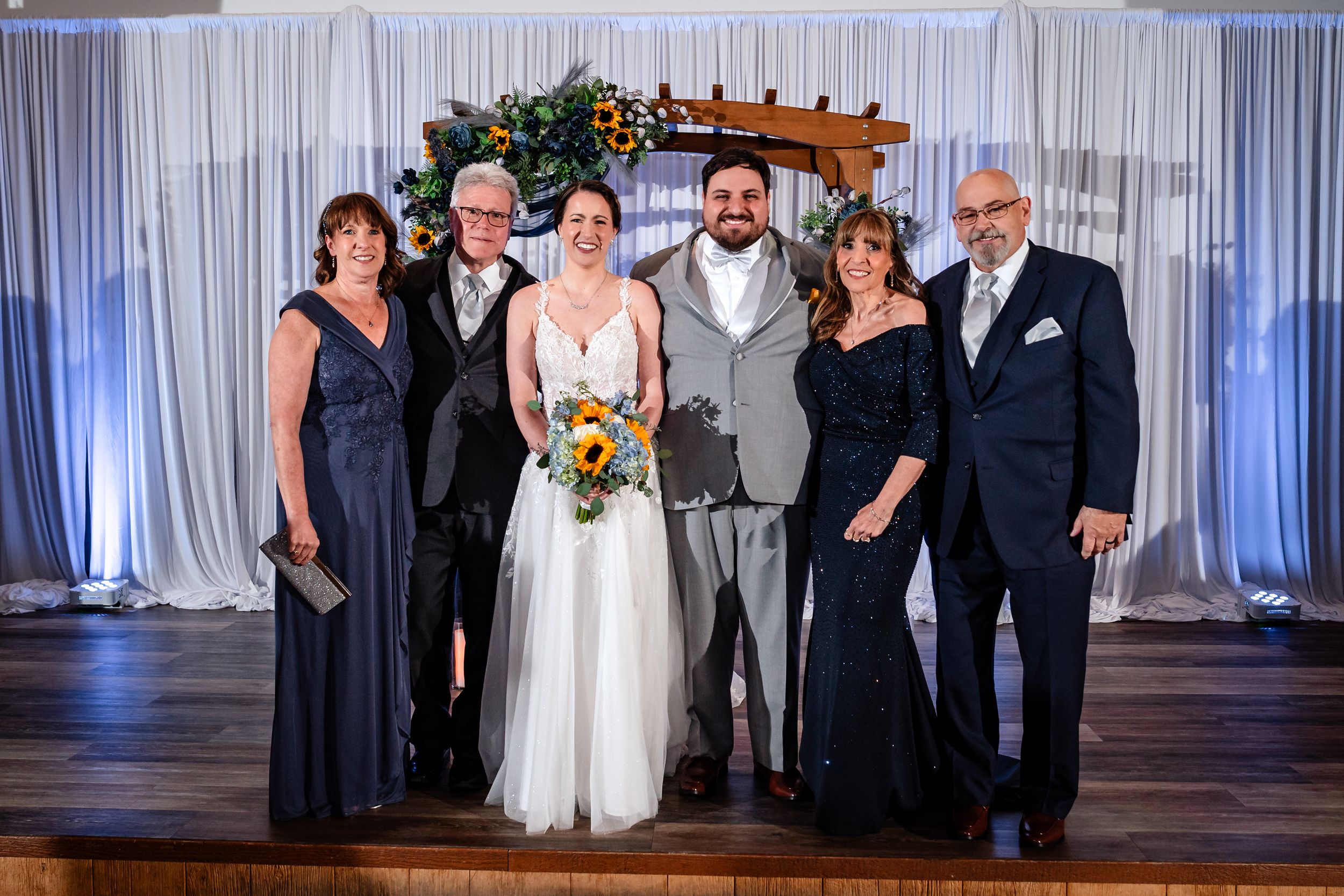 Wedding party of six poses together under floral arch at indoor reception venue.
