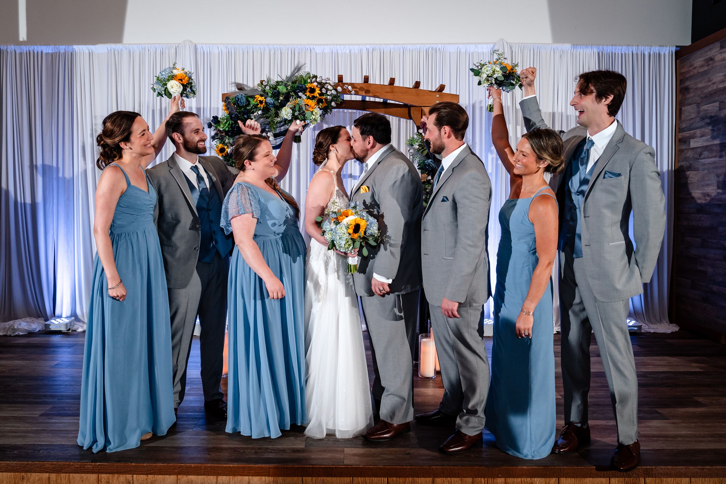 Wedding party in blue and gray attire cheers as bride and groom kiss beneath a floral arch at indoor venue.