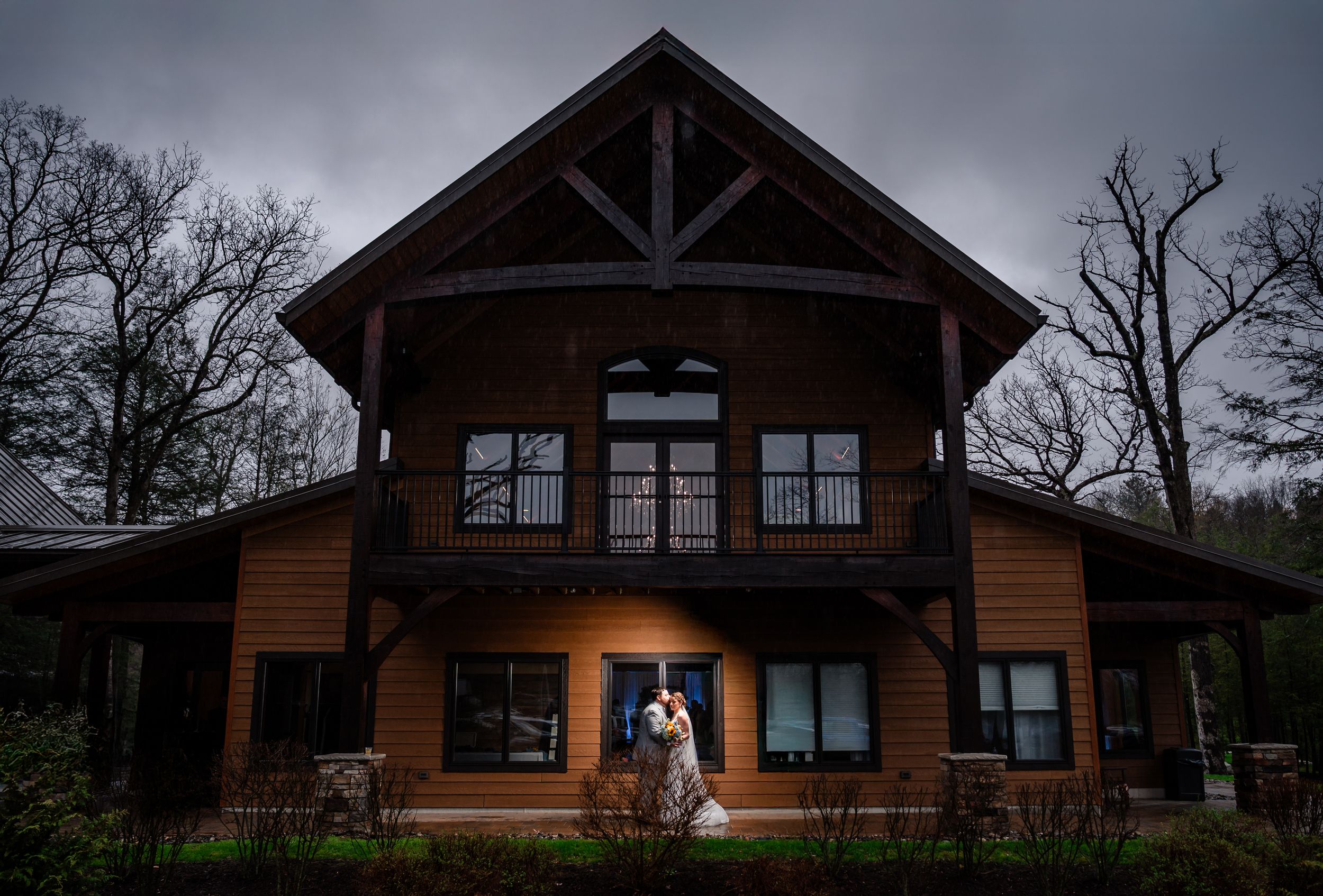 Bride and groom standing outside a rustic wooden lodge venue under dramatic cloudy skies at dusk.