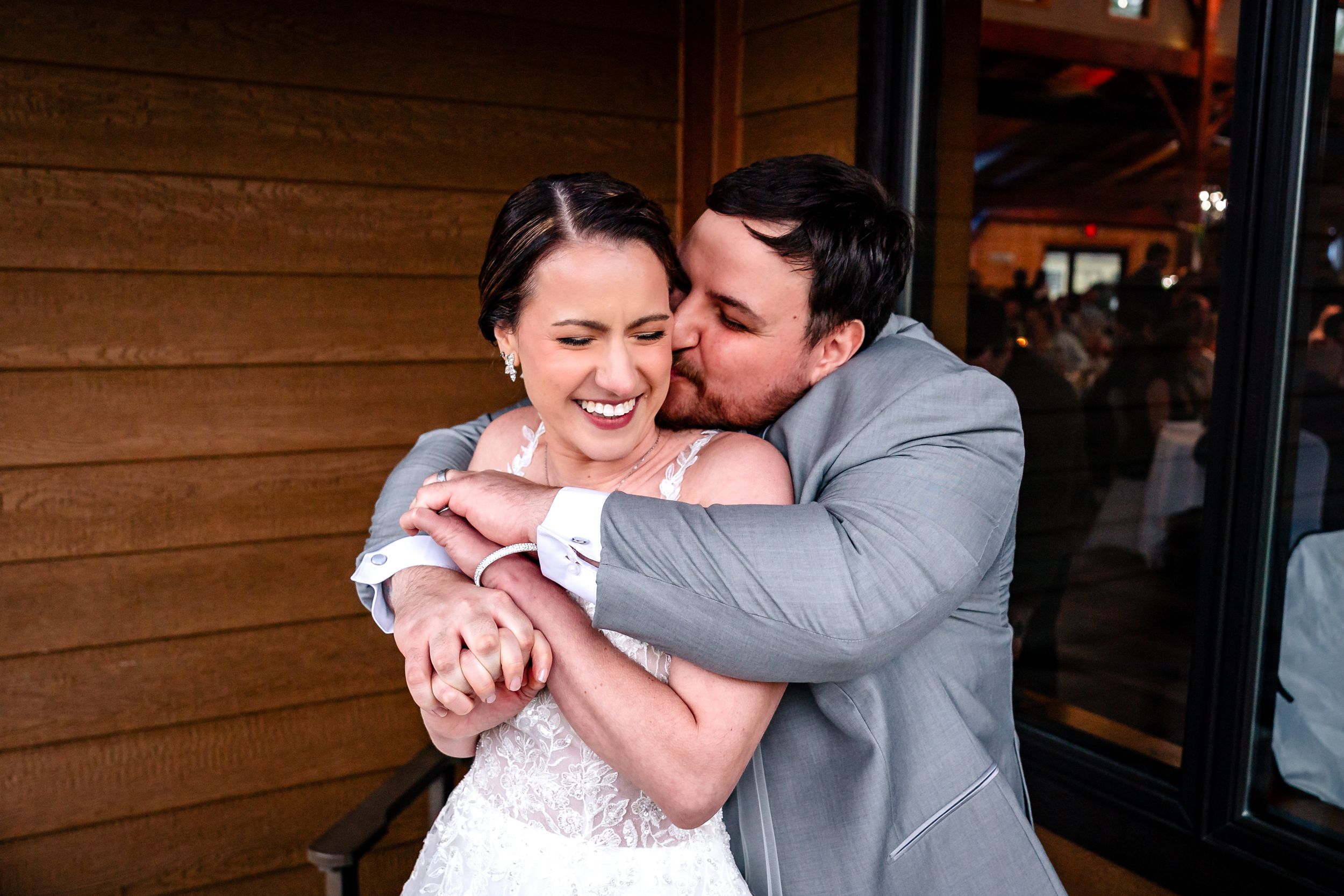 Groom in gray suit hugging and kissing smiling bride on cheek outside rustic wooden wedding venue.