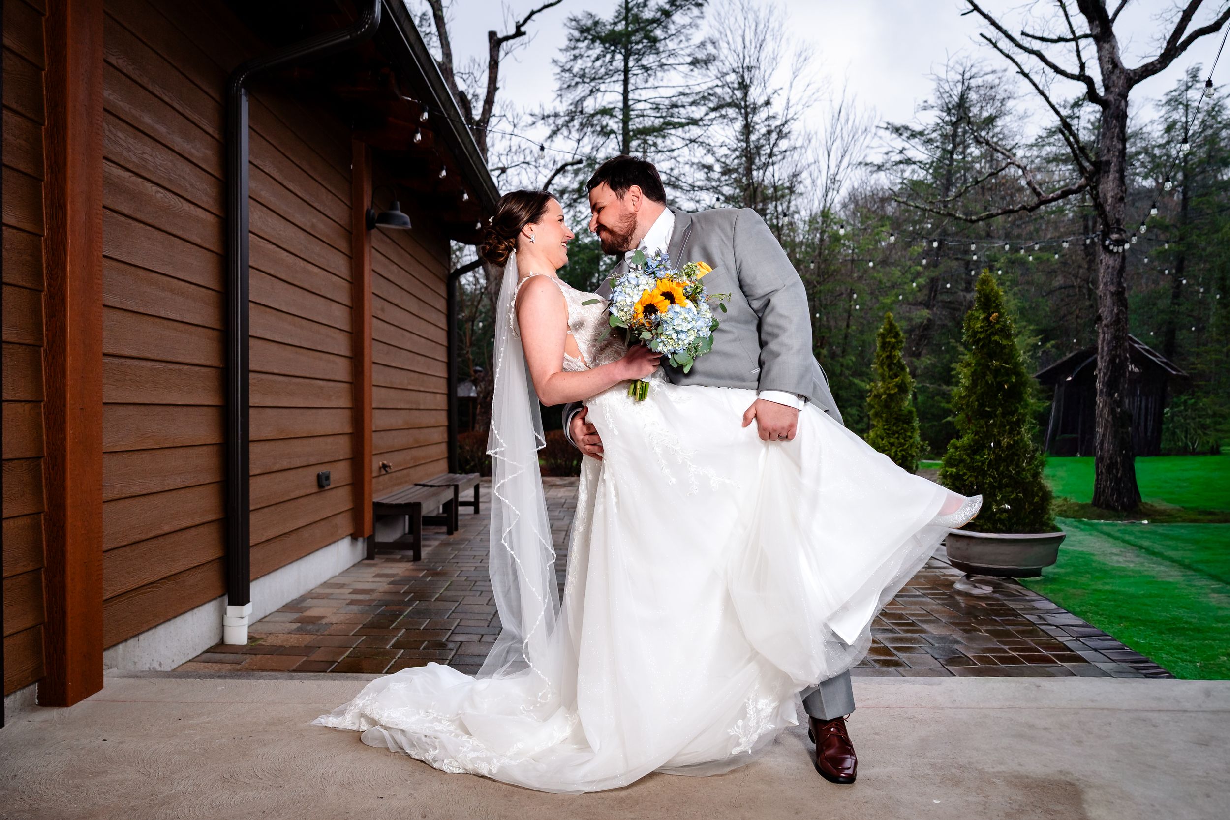 Bride and groom share romantic dip pose outside rustic venue with sunflower bouquet on rainy day.