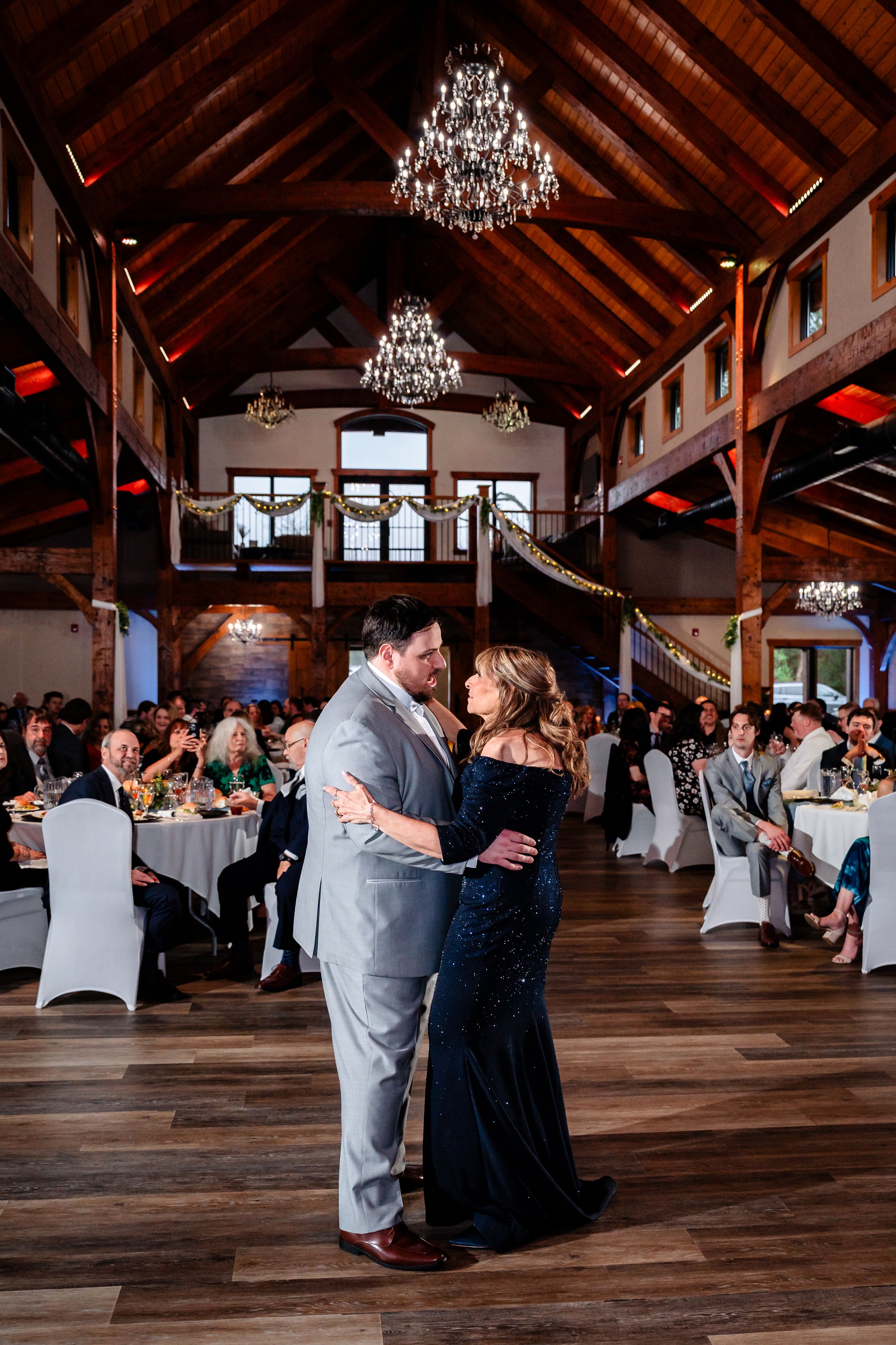 Couple shares first dance in elegant rustic barn wedding venue with chandeliers and guests seated at white-draped tables.
