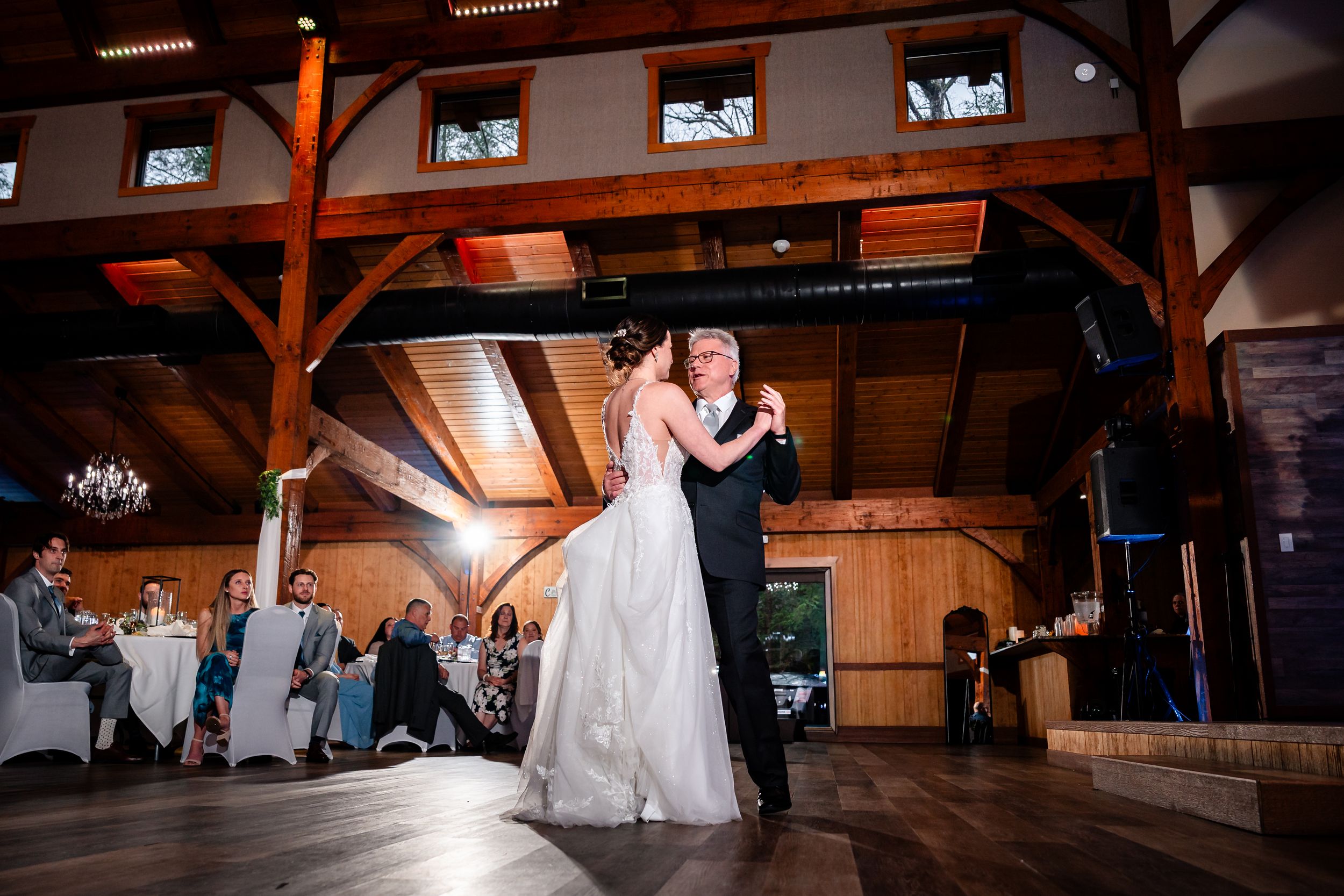 Bride and groom share their first dance in a rustic wooden barn venue with guests watching from surrounding tables.