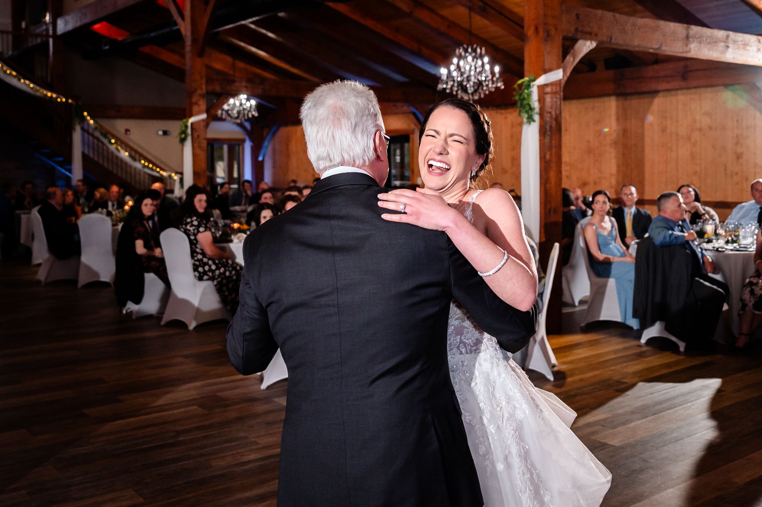 Bride sharing joyful father-daughter dance at rustic wedding reception venue with chandelier lighting.