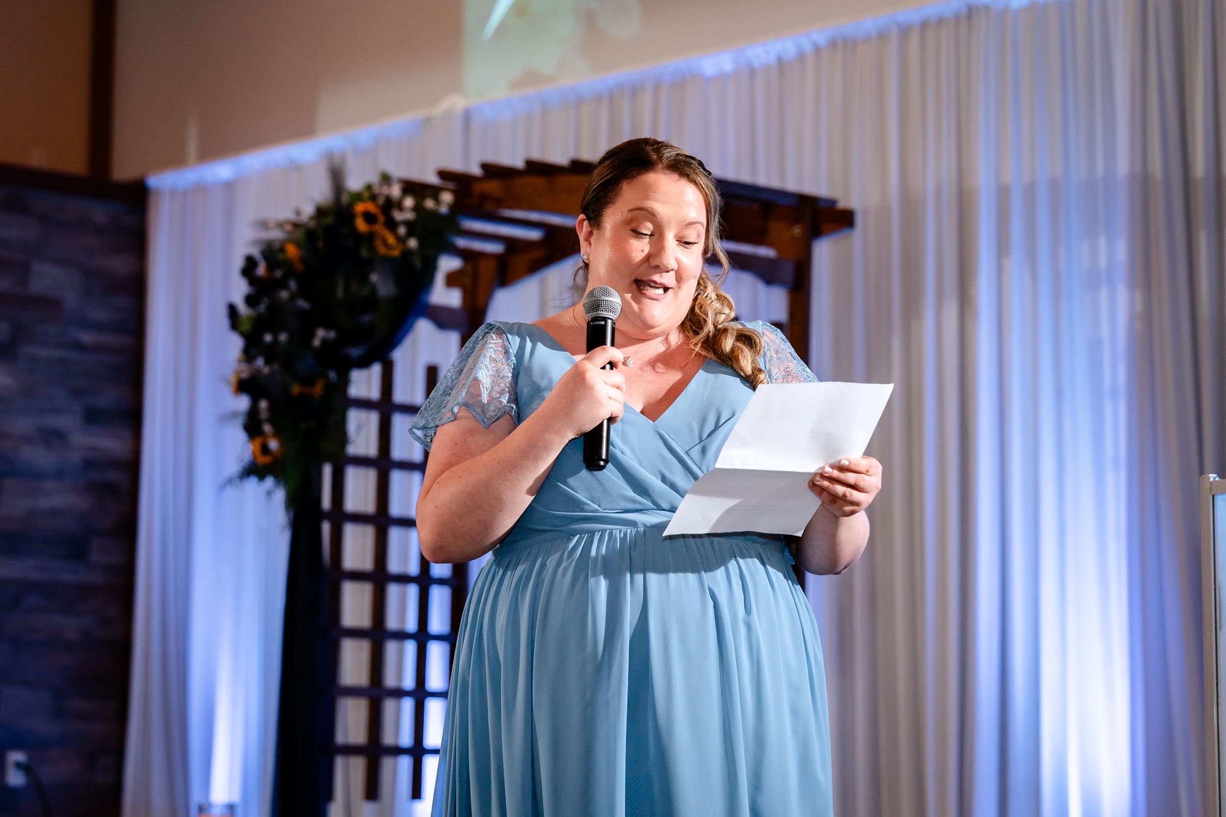 Bridesmaid in blue dress delivers speech at microphone during wedding reception with floral backdrop.