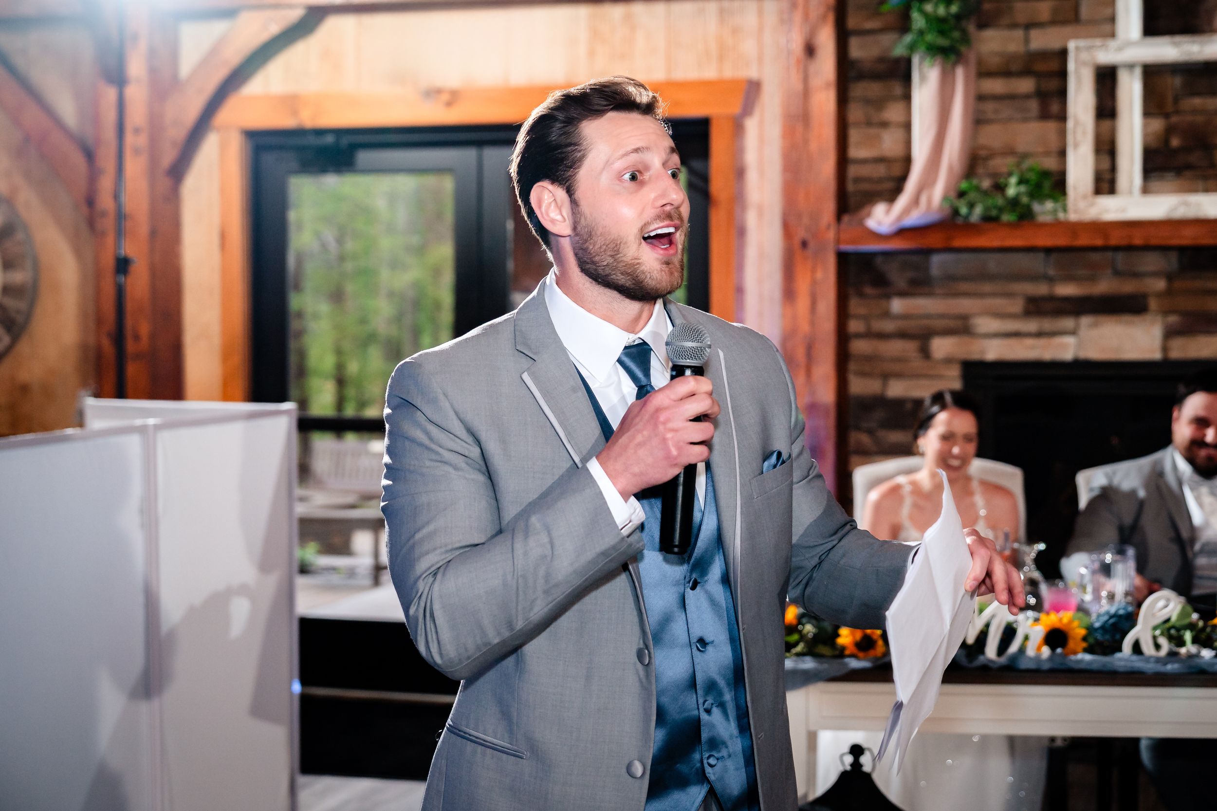 Groomsman in gray suit delivering heartfelt wedding speech with microphone at rustic reception venue.