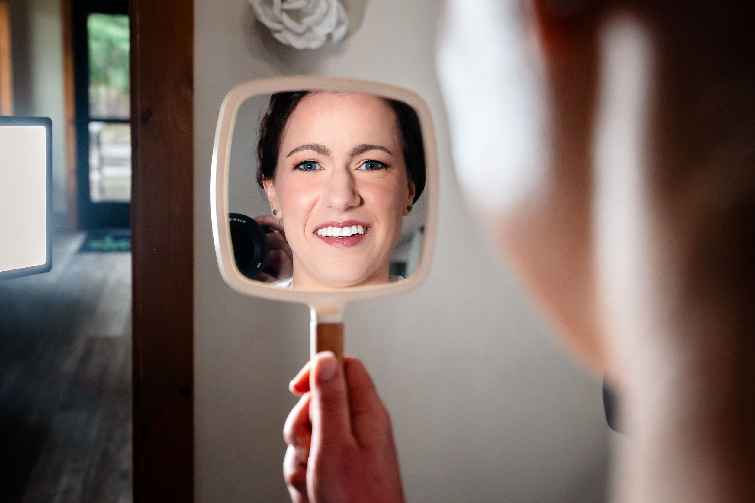 Woman smiling at her reflection in a handheld mirror, captured in a bright indoor setting.