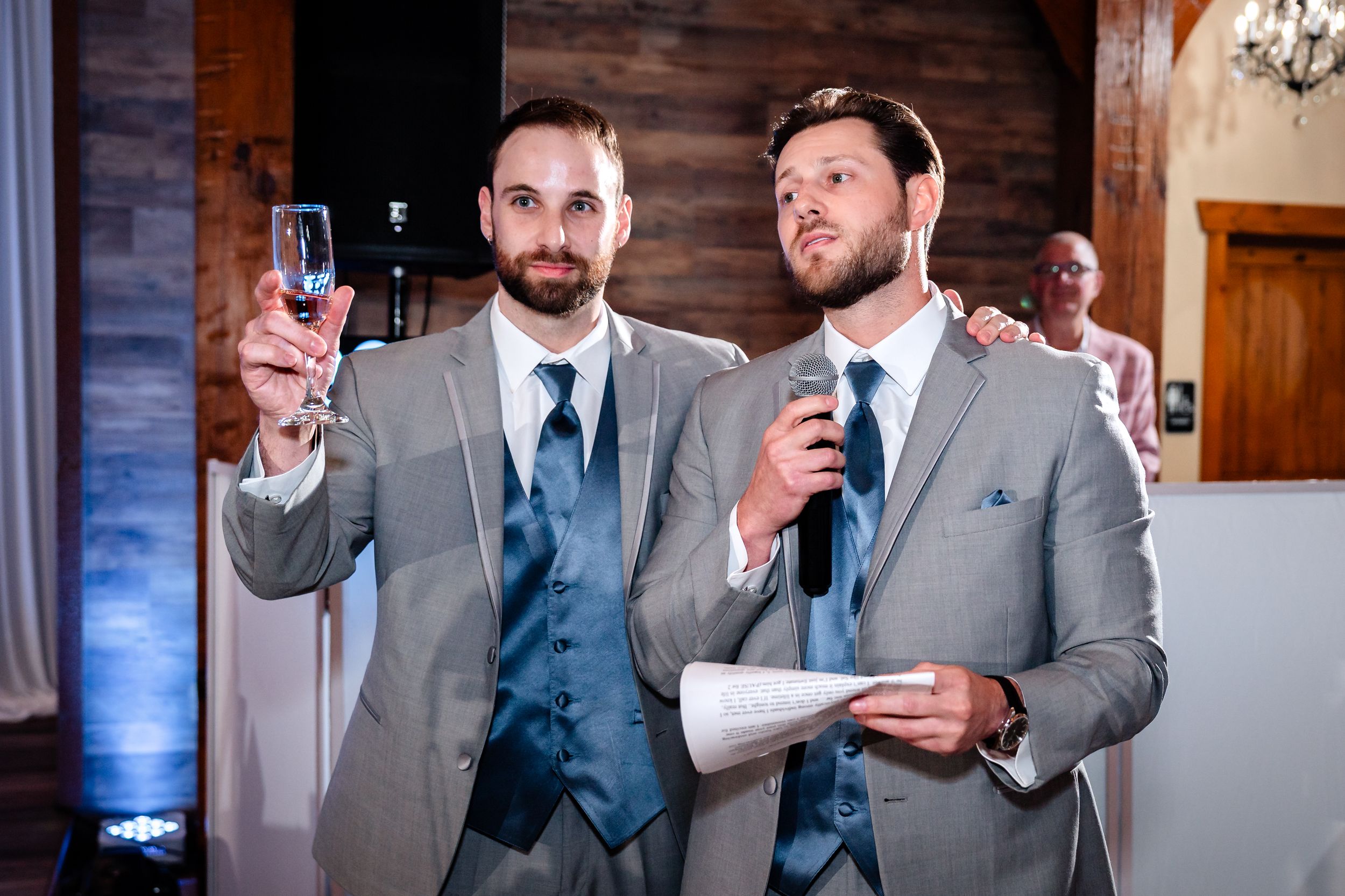 Two groomsmen in grey suits delivering a toast with a champagne glass raised at a wedding reception.