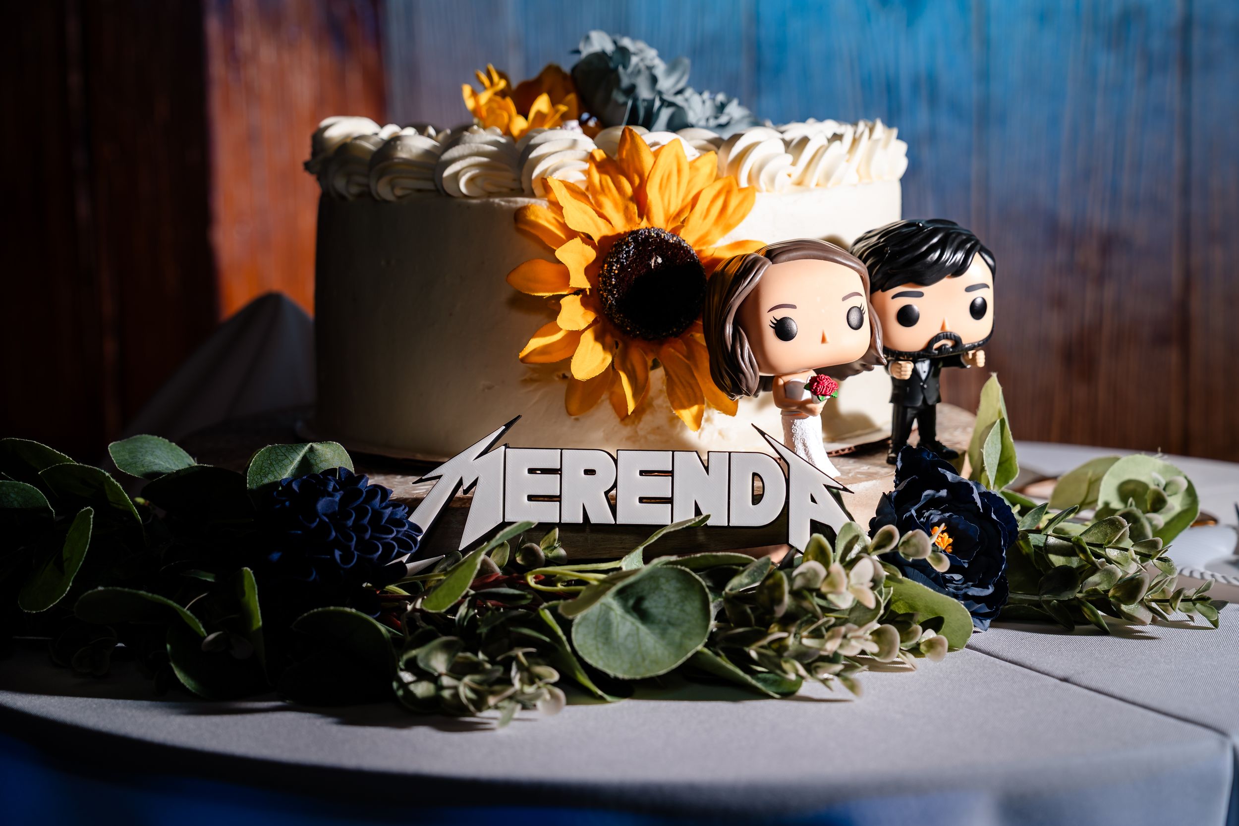 Wedding cake with sunflower topper, Funko Pop bride and groom figurines, and Merende metal sign surrounded by greenery.