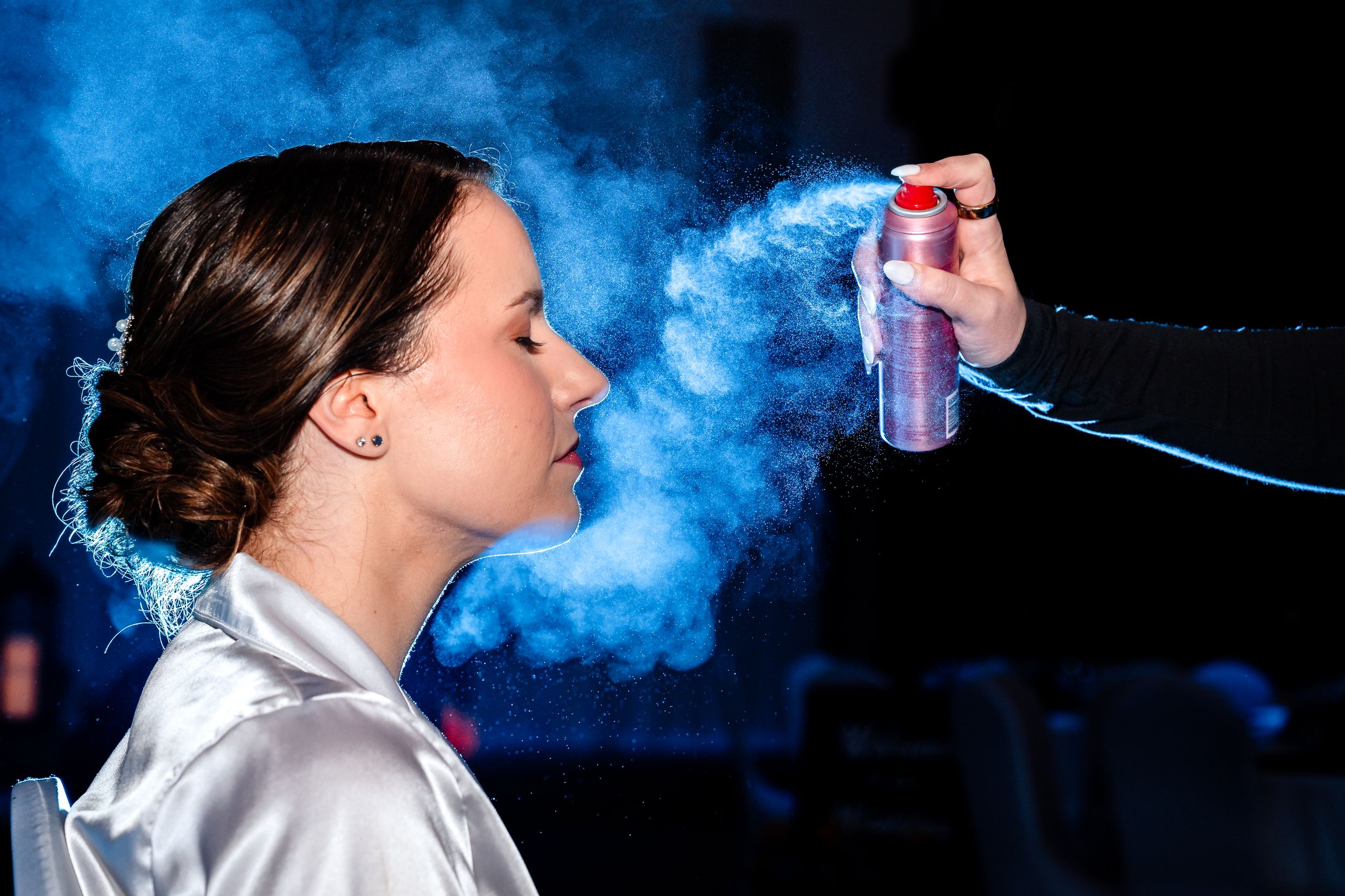 Bride in white robe gets hair sprayed with a cloud of mist during wedding day preparations.