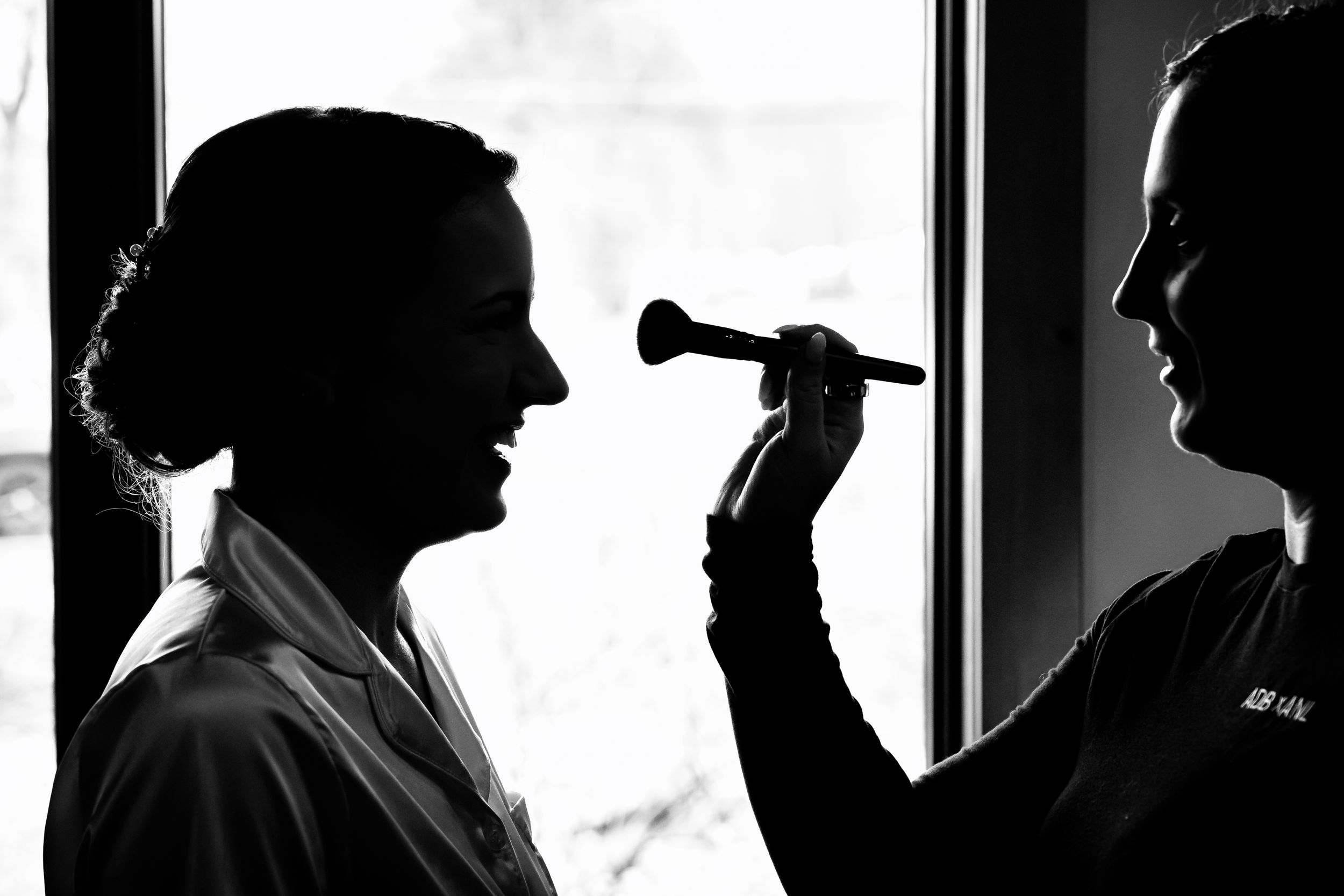 Black and white silhouette of a bride getting makeup applied before her wedding ceremony.