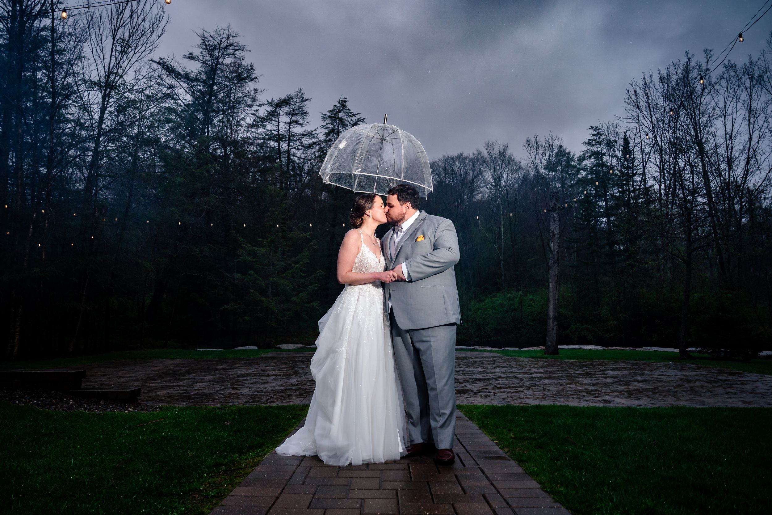 Bride and groom kissing under clear umbrella in rain at dramatic night wedding.
